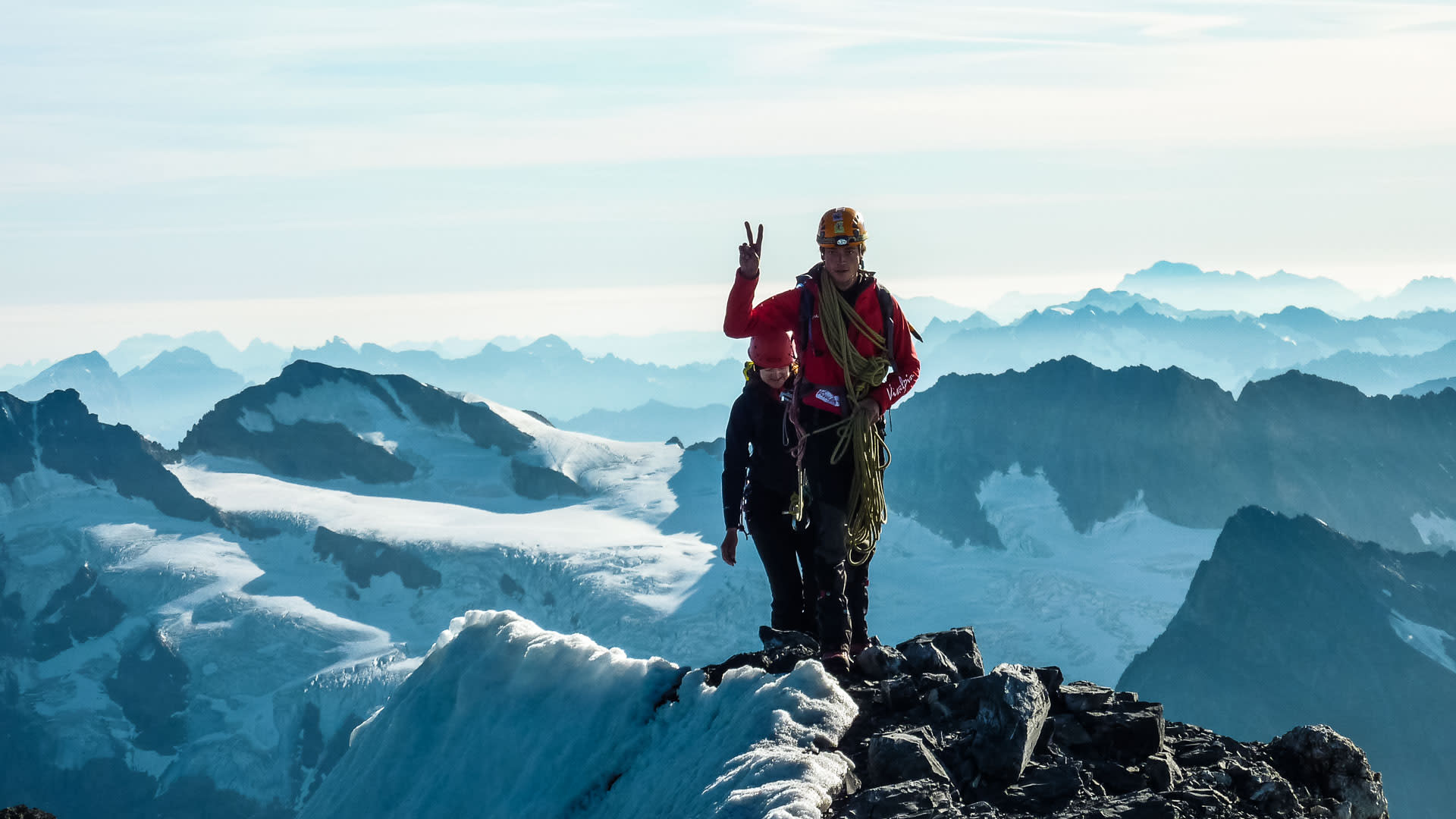 Hochtour auf den Eiger: Bergsteiger mit roter Daunenjacke, Helm und grünem Seil zeigt Victory-Zeichen auf felsigem Gipfelgrat
