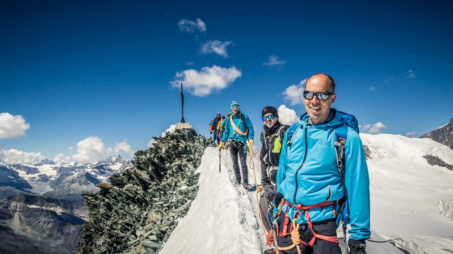 Drei Bergsteiger als Seilschaft auf dem Schneegrat des Allalinhorn mit Gipfelkreuz im Hintergrund.
