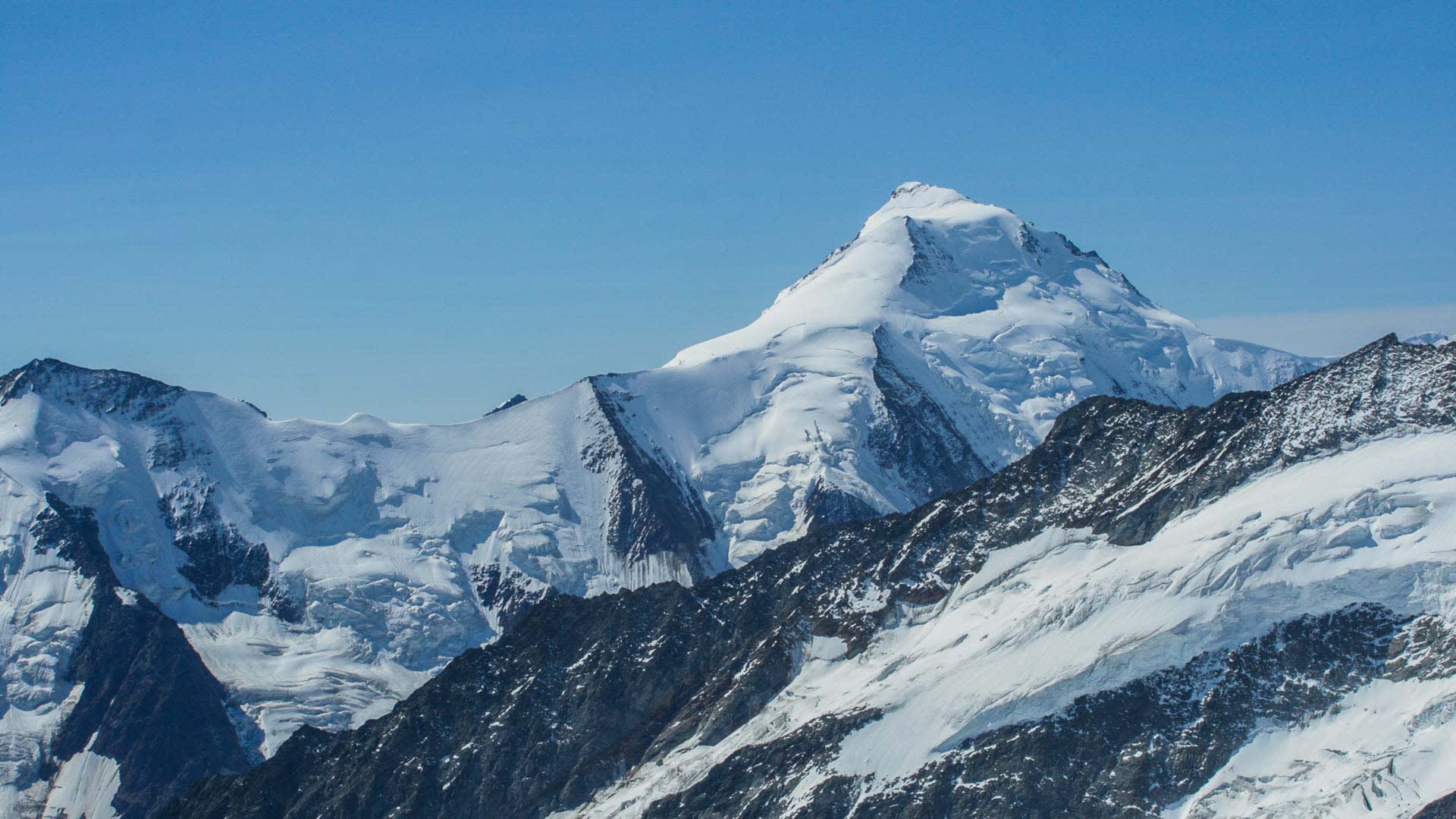 Hochtour im Berner Oberland: Breitbasiger Schneegipfel mit Gletscherflanken, Seracs und dunklen Felsgräten unter wolkenlosem Himmel.