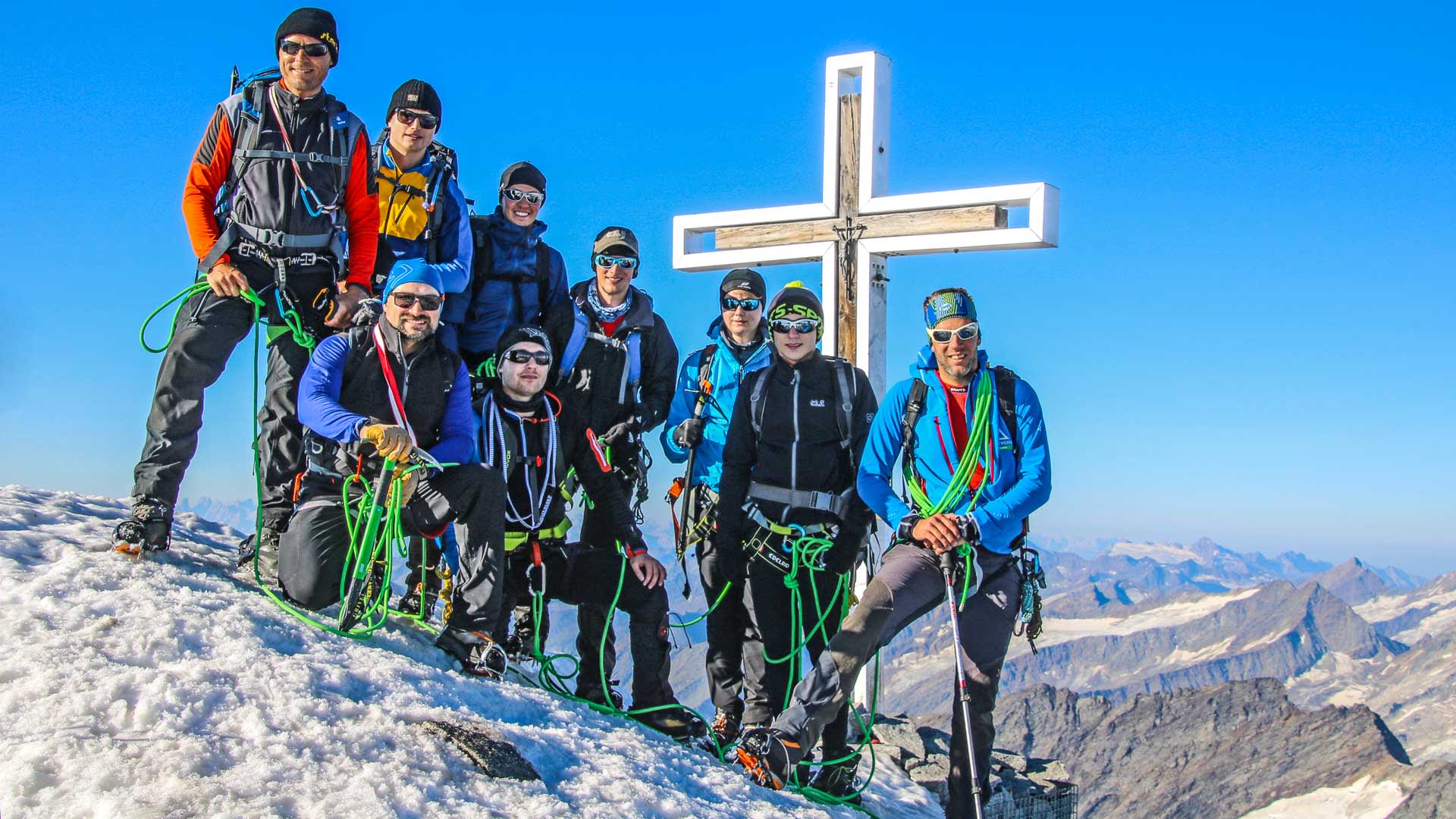 Hochtour auf den Großvenediger: neun Bergsteiger mit Klettergurten und Seilen posieren am weißen Holzgipfelkreuz.