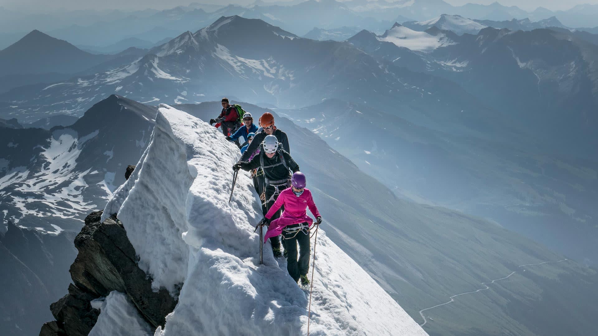 Fünf Bergsteiger traversieren am verschneiten Gipfelgrat des Großglockner mit Seil, Helmen und Klettergurten gesichert.