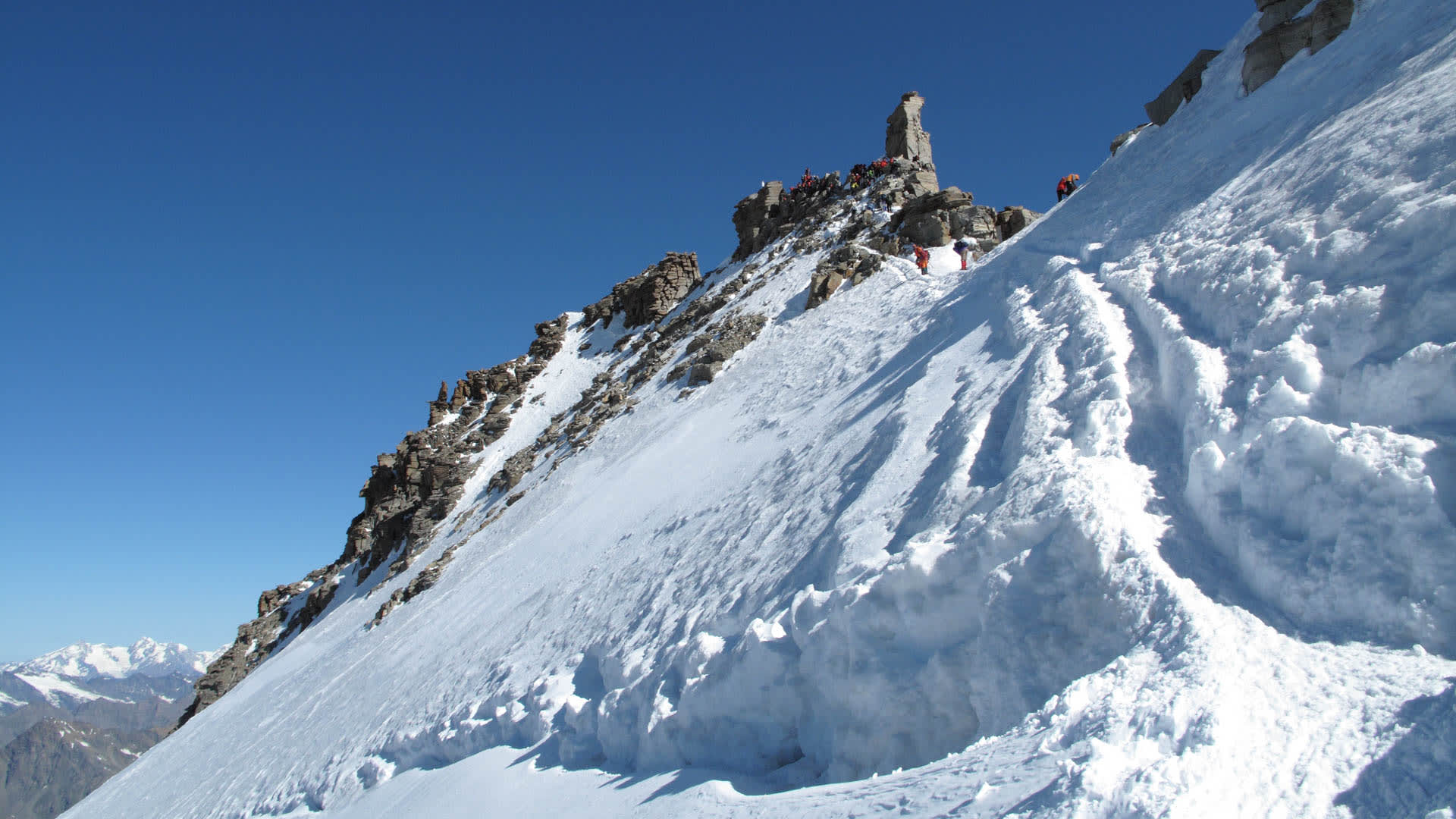 Hochtour Gran Paradiso: Bergsteigergruppe am felsigen Gipfelgrat über steilem Schneehang mit Spurrillen und Ferngipfeln
