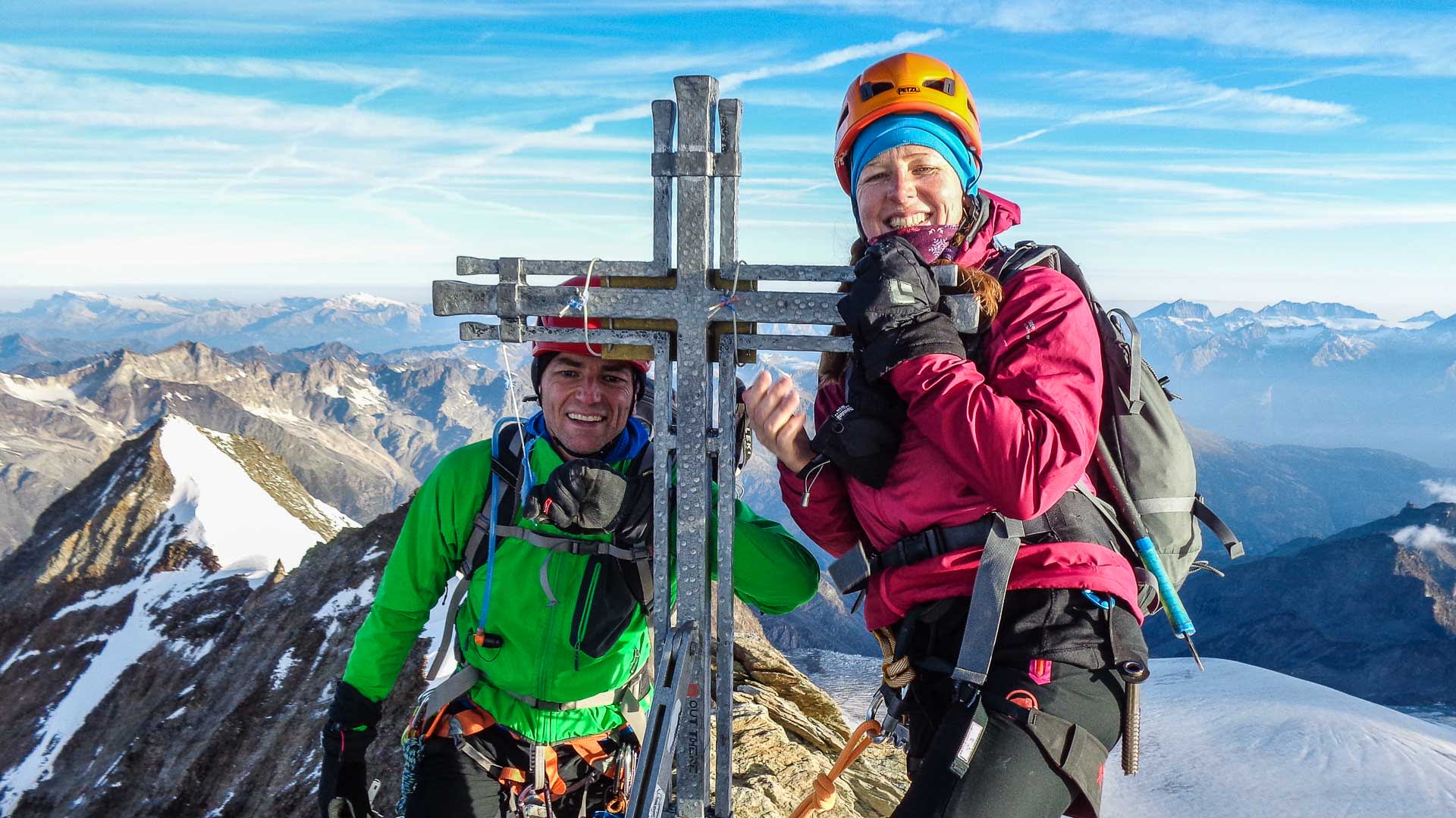 Zwei Bergsteiger am Gipfelkreuz bei einer Hochtour am Nadelgrat mit Hochgebirgspanorama im Hintergrund.
