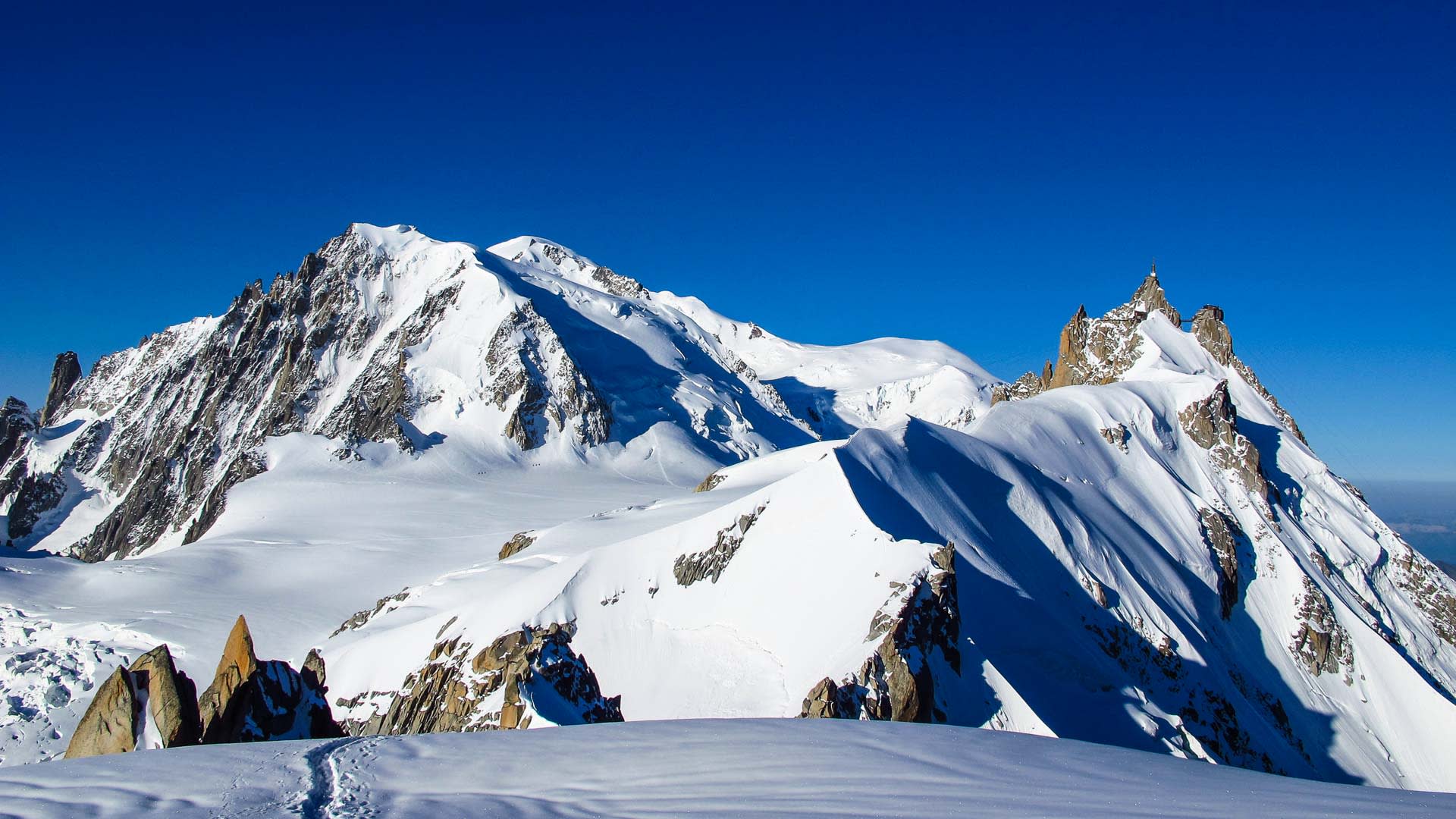 Weitblick auf den schneebedeckten Mont-Blanc-Gipfel mit Gletscherplateau und kleinen Bergsteigern im Vordergrund.