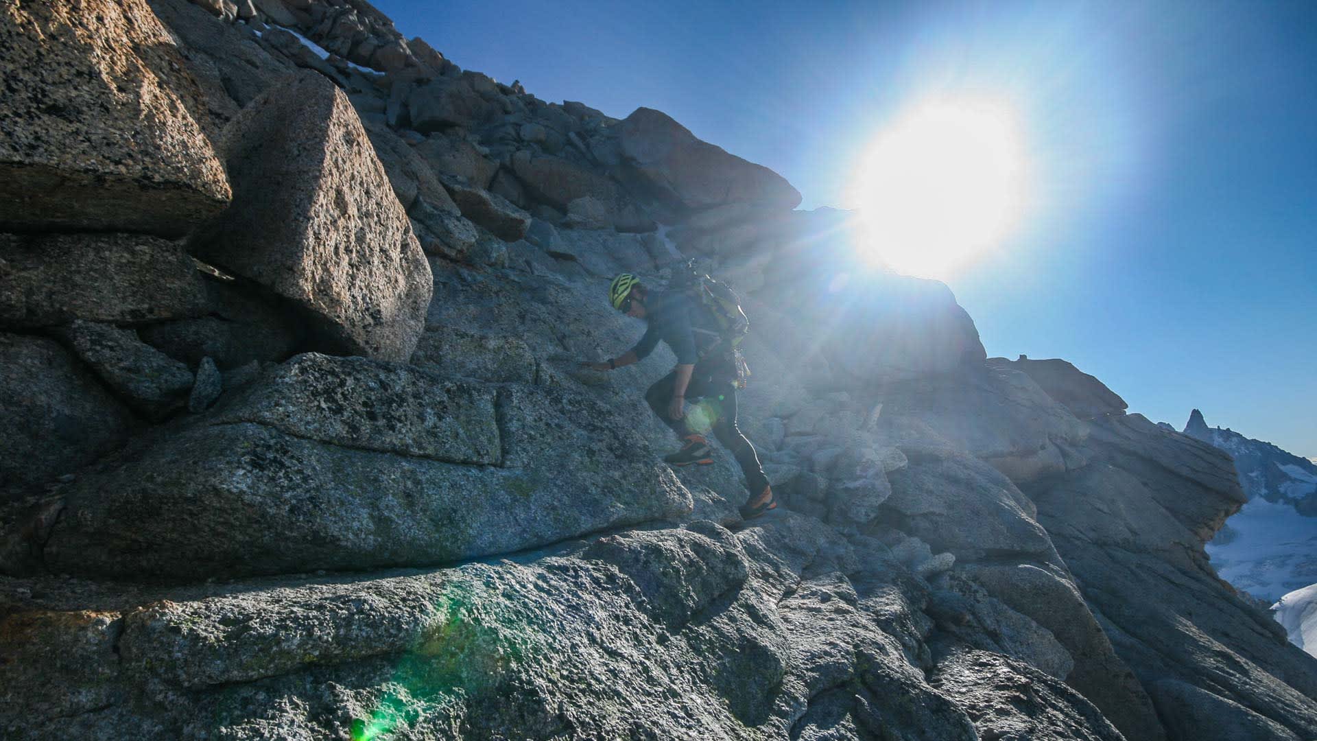 Hochtour in den Alpen: Bergsteiger klettert mit Helm und Rucksack über strukturierten Granitgrat in Froschperspektive.