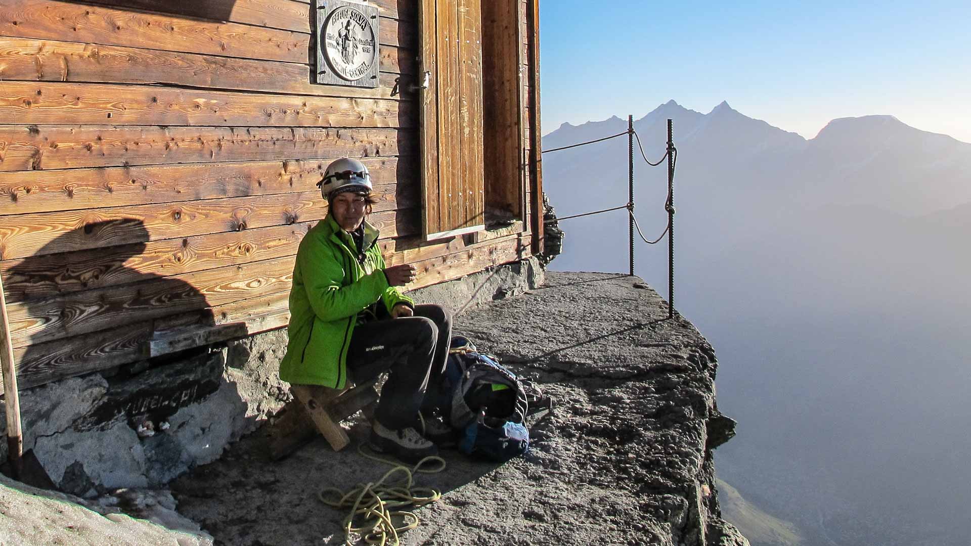 Bergsteiger rastet auf Felsvorsprung an Berghütte bei der Hochtour in Zermatt mit Rucksack und Seil.