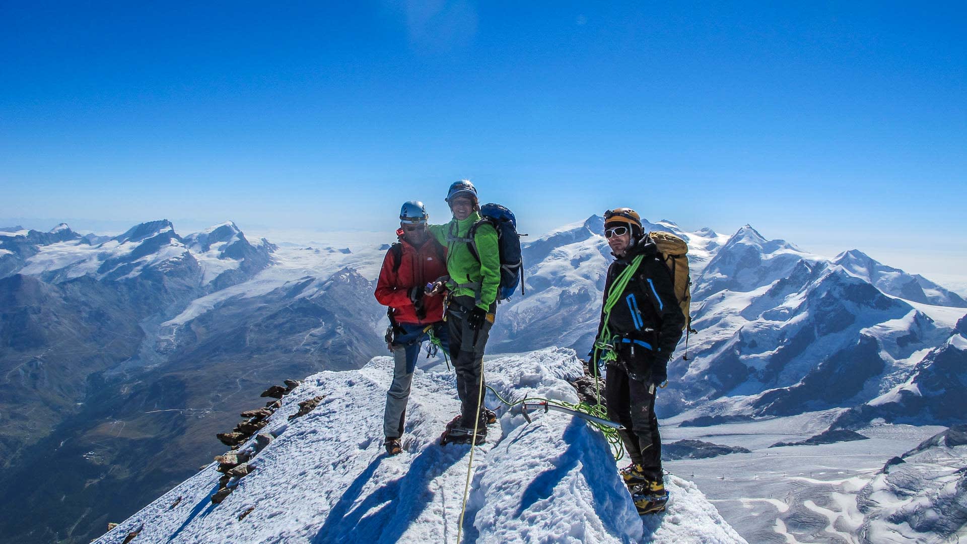 Hochtour Matterhorn: Drei Bergsteiger mit Helm, Steigeisen und grünem Seil stehen auf dem schneebedeckten Gipfelgrat