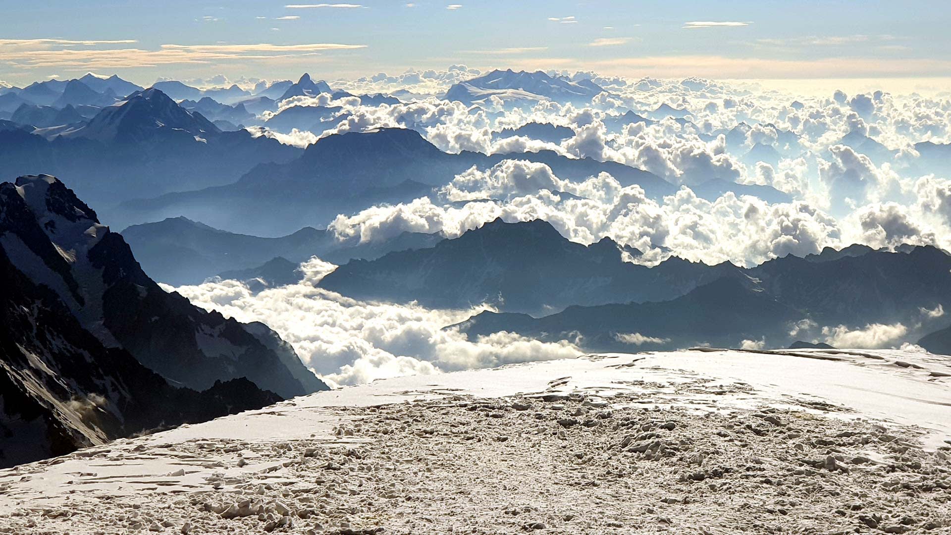 Hochtour Mont Blanc: Blick vom Gipfelbereich über eine Firn­fläche auf Cumulus-Wolkenfelder und aus der Wolkendecke ragende