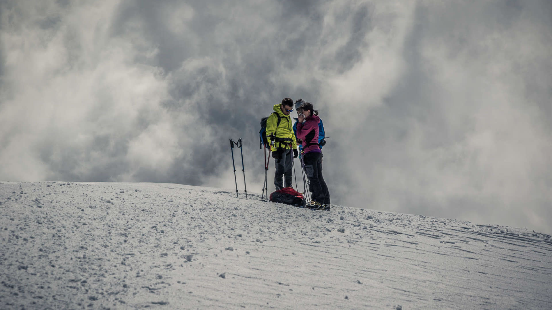 Zwei Bergsteiger rasten mit Trekkingstöcken auf einem weiten Schneefeld im Wallis bei bewölktem Himmel.