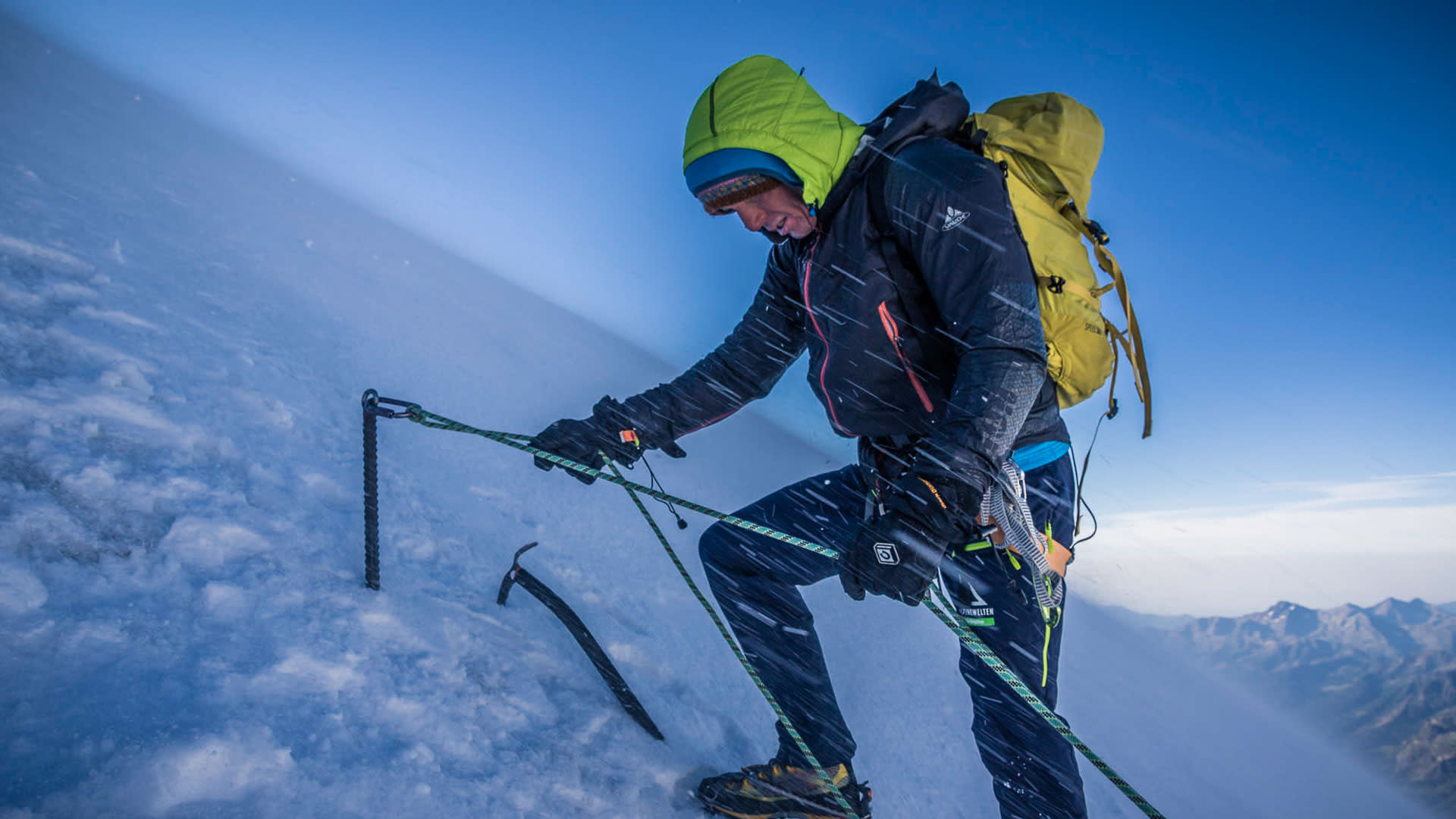 Hochtour im Wallis: Bergführer sichert mit Seil und Eisenanker an steilem Schneehang, Steigeisen und Pickel sichtbar.