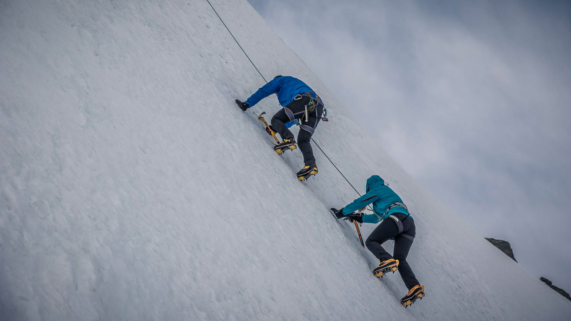 Zwei Bergsteiger klettern mit Steigeisen und Pickel am Seil eine steile Schneeflanke in den Walliser Alpen aufwärts.