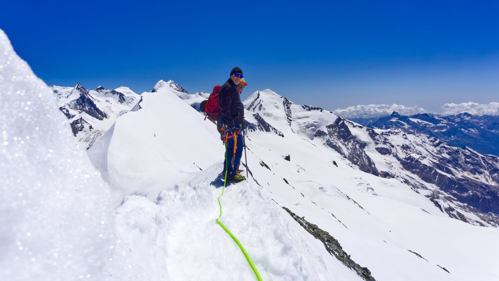 Zwei Bergsteiger bei einer Hochtour im Wallis stehen auf einem schmalen Schneegrat, verbunden durch ein neongrünes Seil