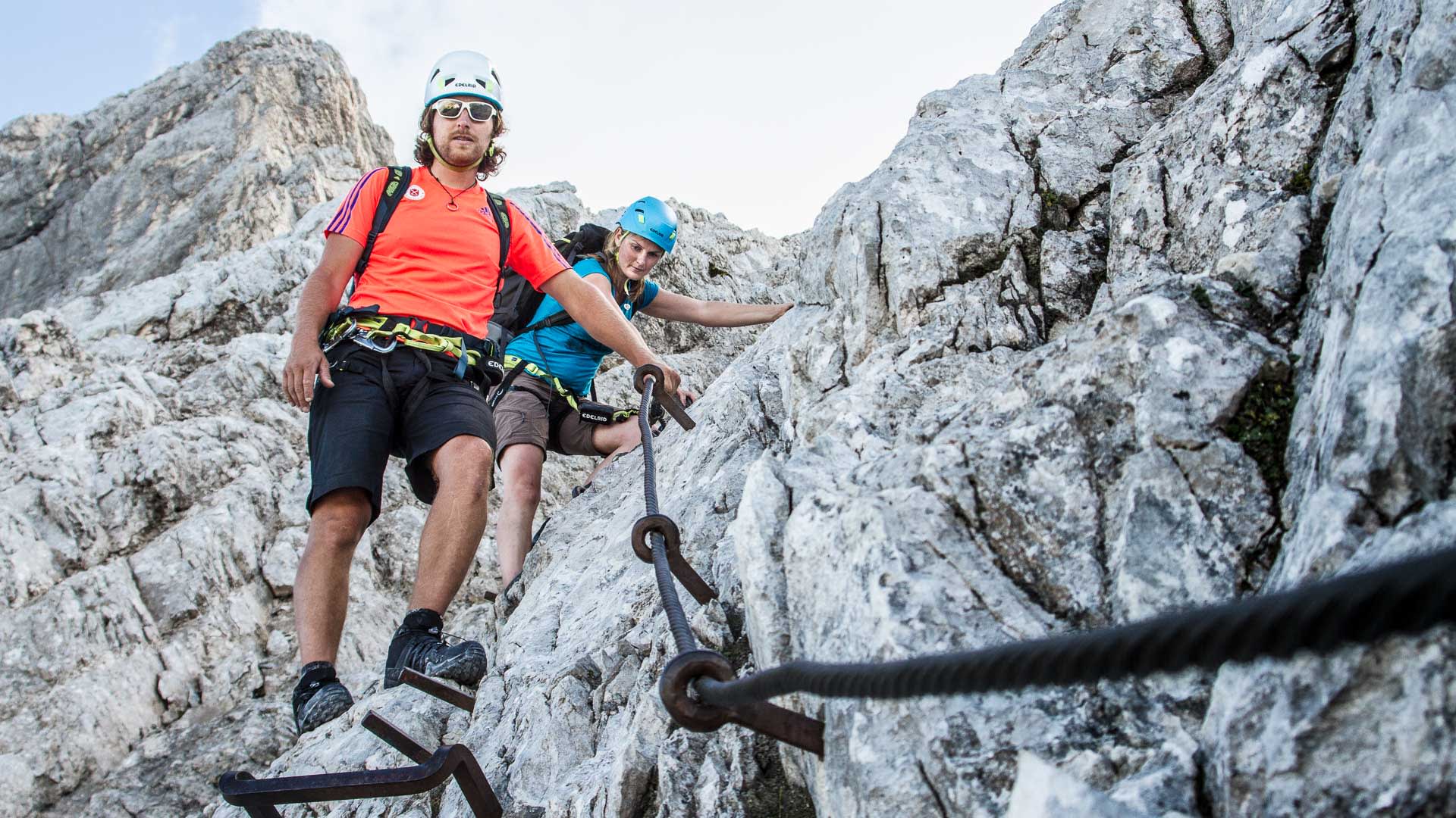 Zwei Kletterer steigen am Klettersteig der Alpspitze ab, gesichert an Fixseil und eisernen Griffbügeln im Kalkfels.