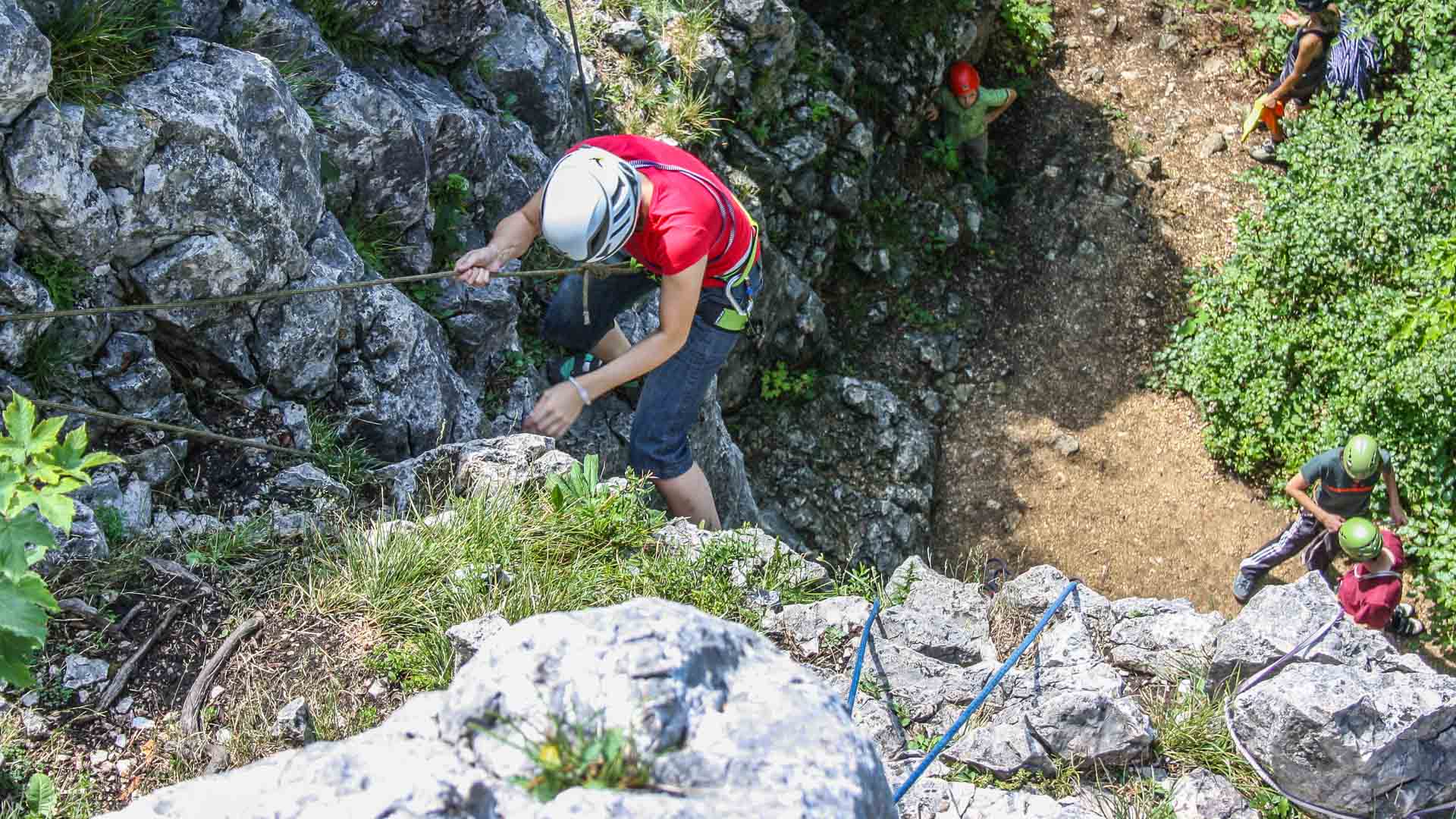 Vogelperspektive auf Klettergruppe der Alpinschule an hellem Kalkfelsen: Personen mit Helmen und Gurten sichern sich gegenseitig