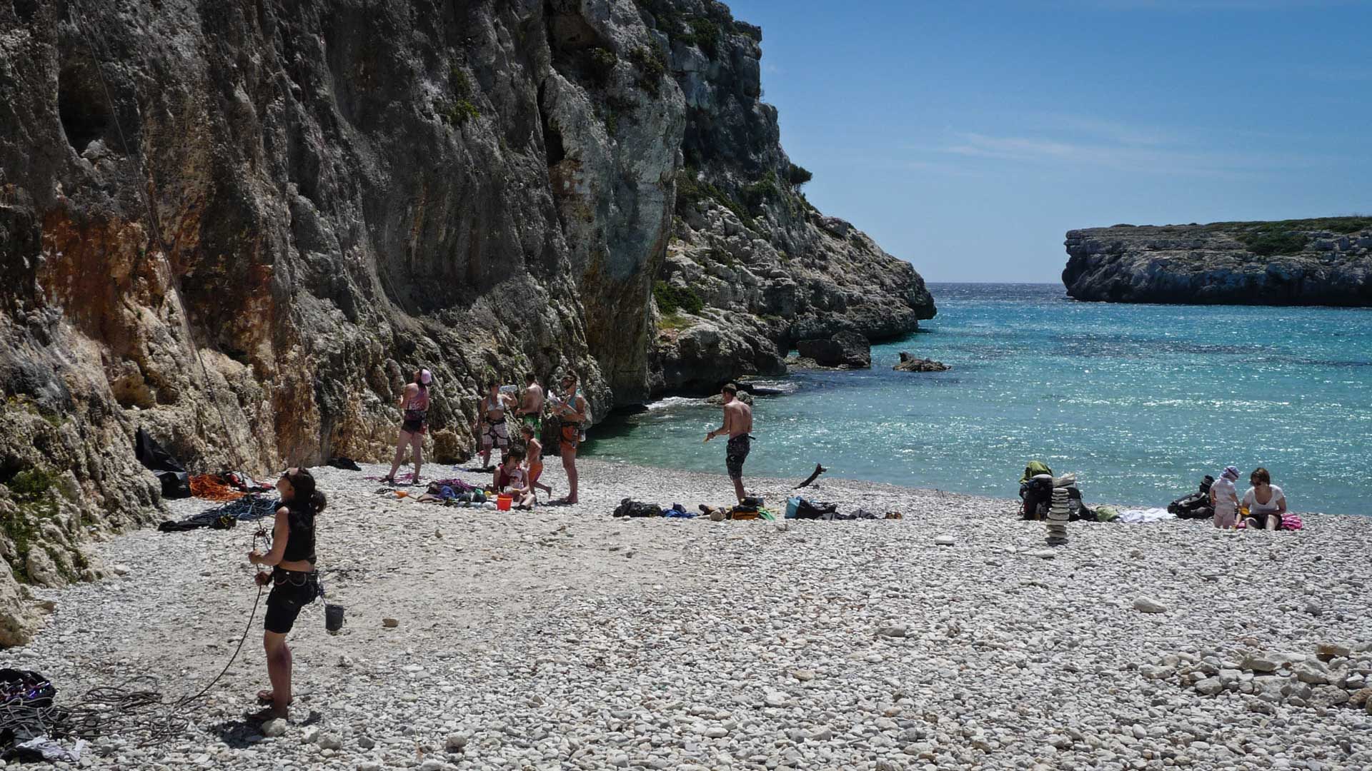Klettergruppe mit Ausrüstung am Kiesstrand der Cala Magraner auf Mallorca vor steil aufragender Kalkfelswand.