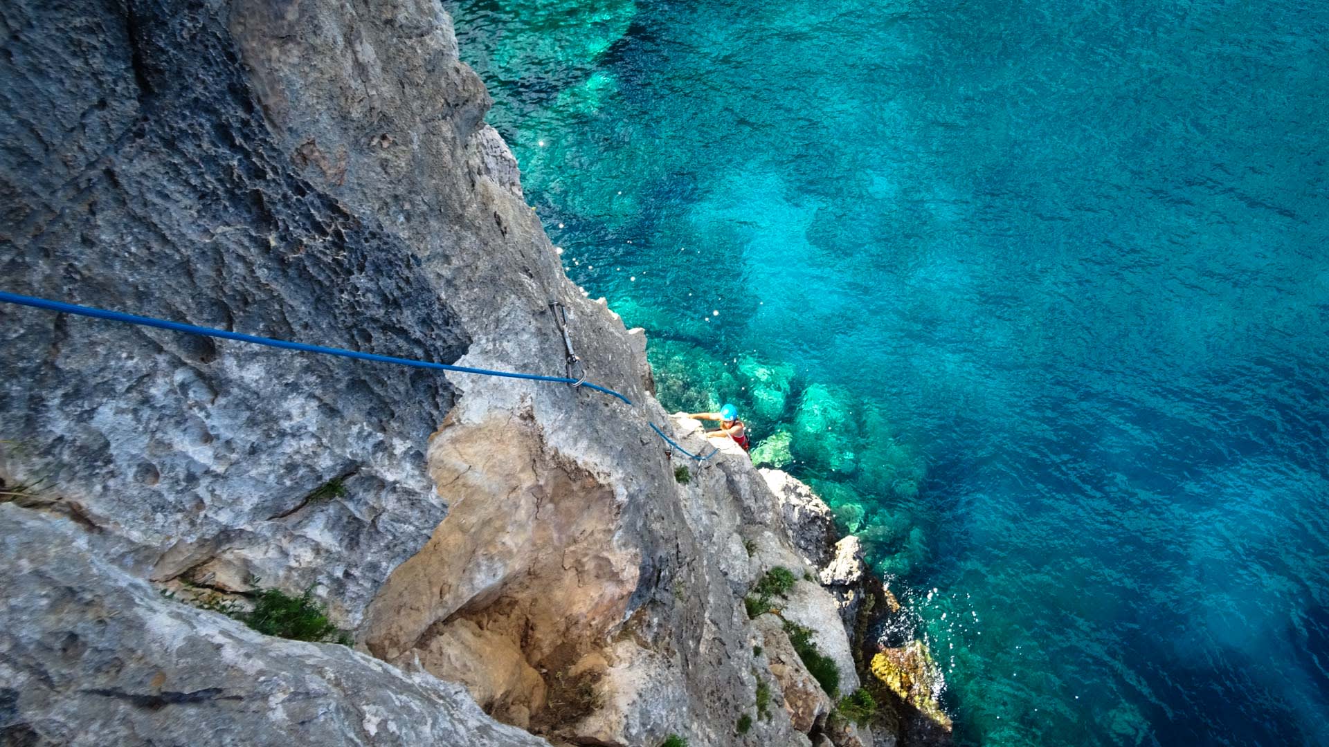 Kletterer mit blauem Helm und roter Jacke klettert an grauer Felswand direkt über türkisblauem Meerwasser in Sardinien.