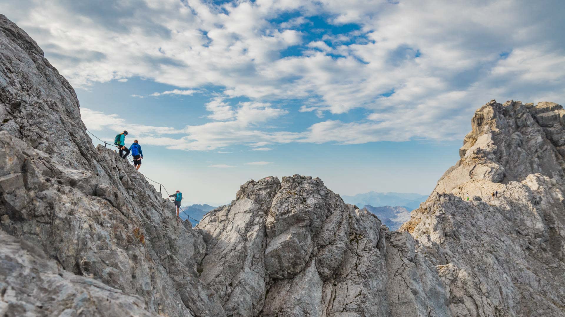 Klettern am Watzmann: drei Kletterer traversieren am Fixseil entlang steilem Kalkstein-Gratkamm, blauer Himmel mit Quellwolken.