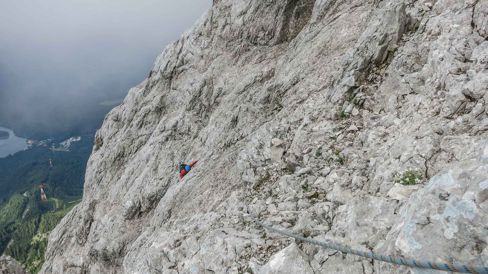 Klettern an der Zugspitze: Kletterer mit blauem Helm und roter Jacke traversiert Kalkfelsband, Fixseil im Vordergrund.