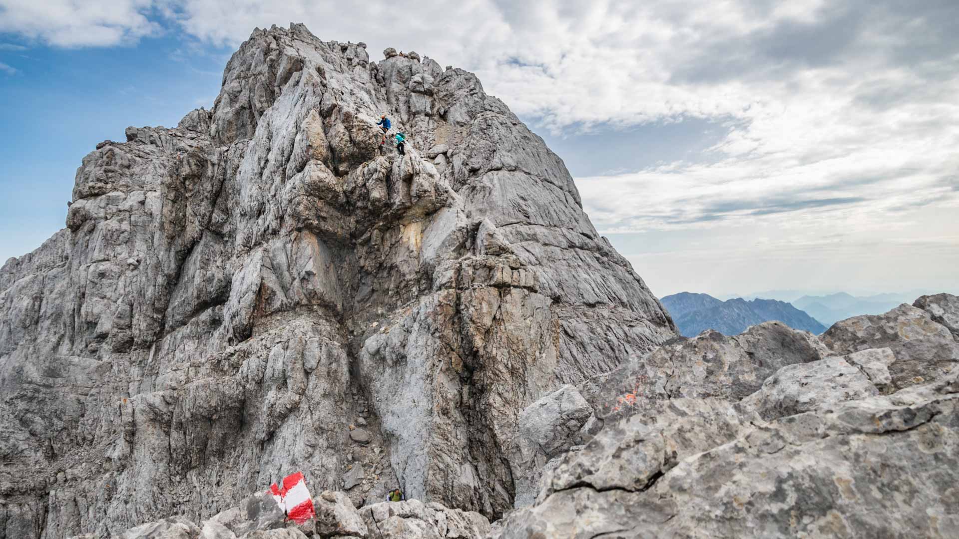Zwei Kletterer steigen in blauer und türkiser Jacke am steil aufragenden Kalkfelsen-Gipfelturm des Watzmann auf.