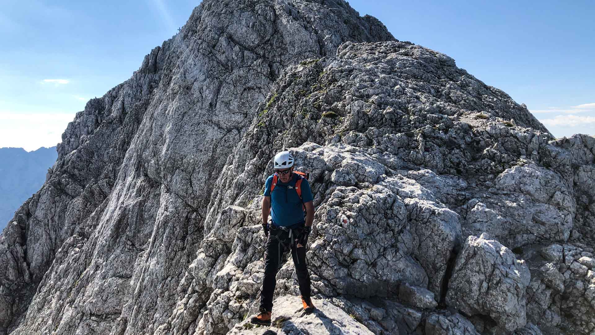 Kletterer mit Helm und Gurt steht auf felsigem Grat am Hochkalter, Schöner Fleck im Hintergrund.