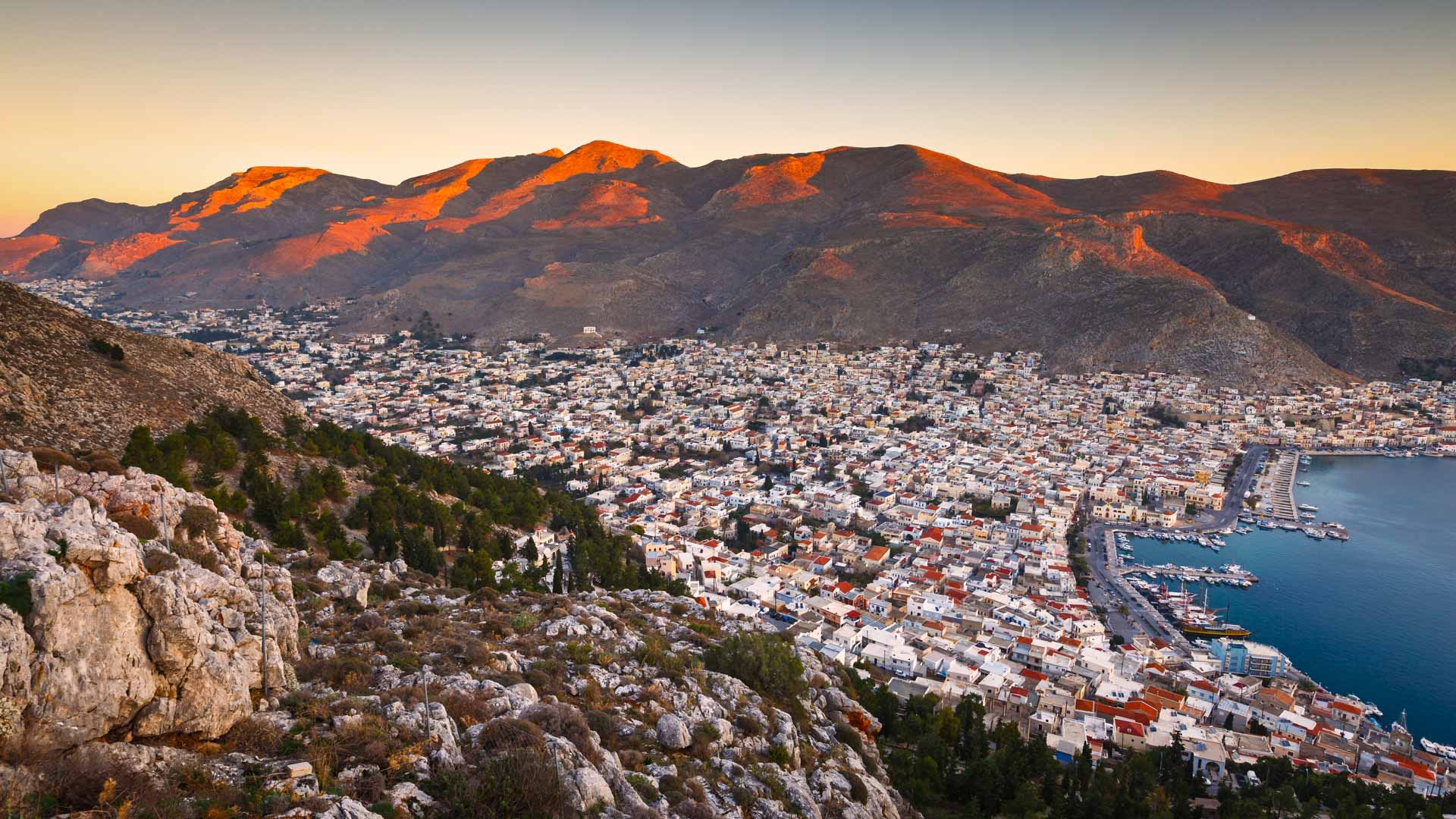 Klettern auf Kalymnos: Panorama der Hafenstadt im Talkessel mit felsigen Kalksteinbergen, die beim Sonnenuntergang orangerot