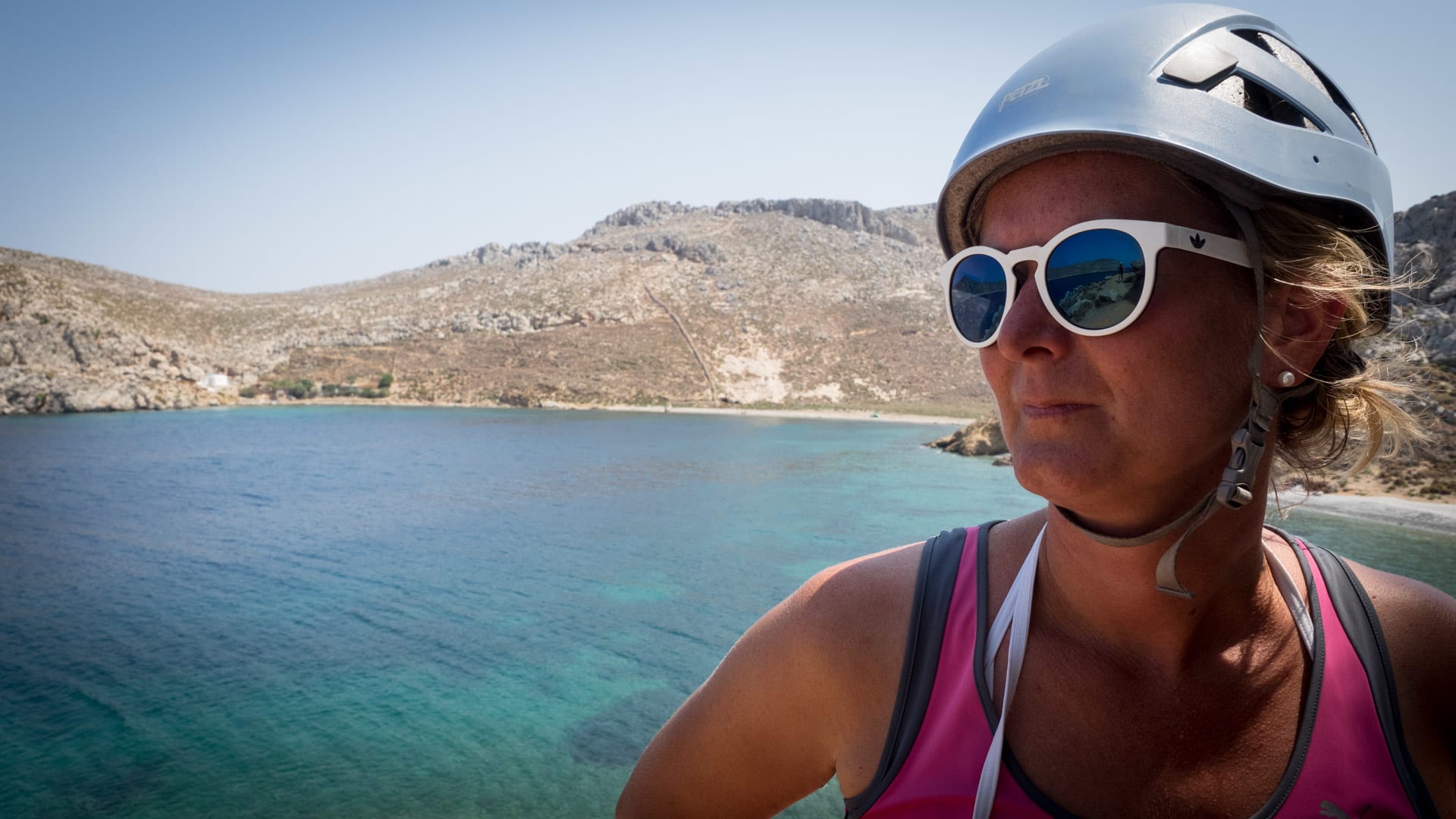 Kletterin mit Petzl-Helm und Klettergeschirr vor türkisfarbener Bucht und trockenem Felsküstenpanorama auf Kalymnos.