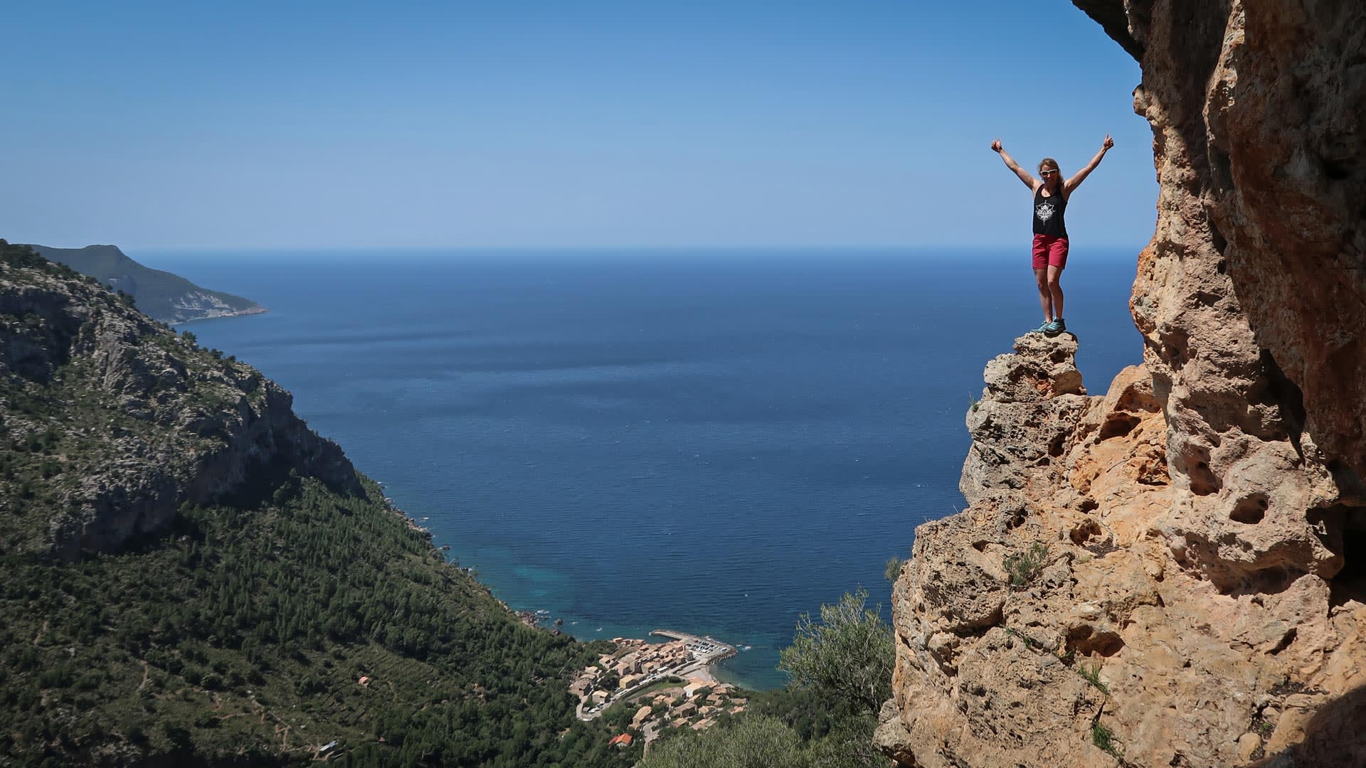 Kletterer steht mit erhobenen Armen auf verwittertem Tafoni-Kalkfelsturm beim Klettern Mallorca, im Hintergrund Küstensiedlung