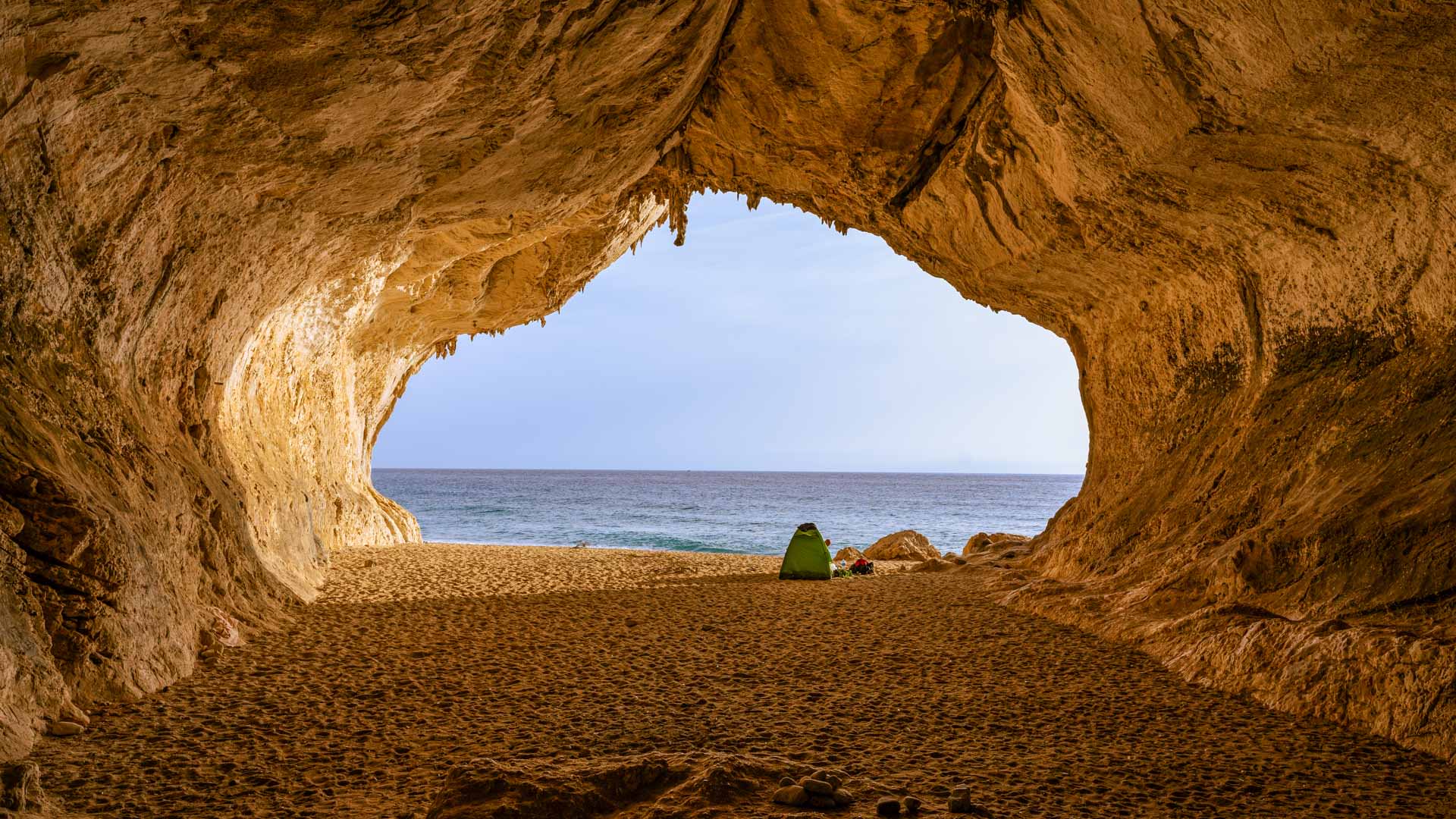 Kletterreise Sardinien: Blick aus einer Meereshöhle aus orangefarbenem Kalkstein auf Sandstrand und türkisblaues Meer