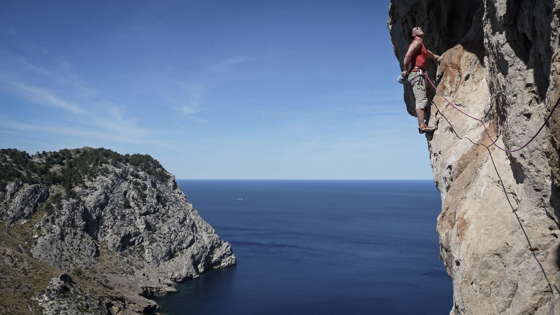 Kletterer an senkrechter Kalksteinwand auf Mallorca mit Blick auf tiefblaue Meeresbucht und felsige Halbinsel im Hintergrund.