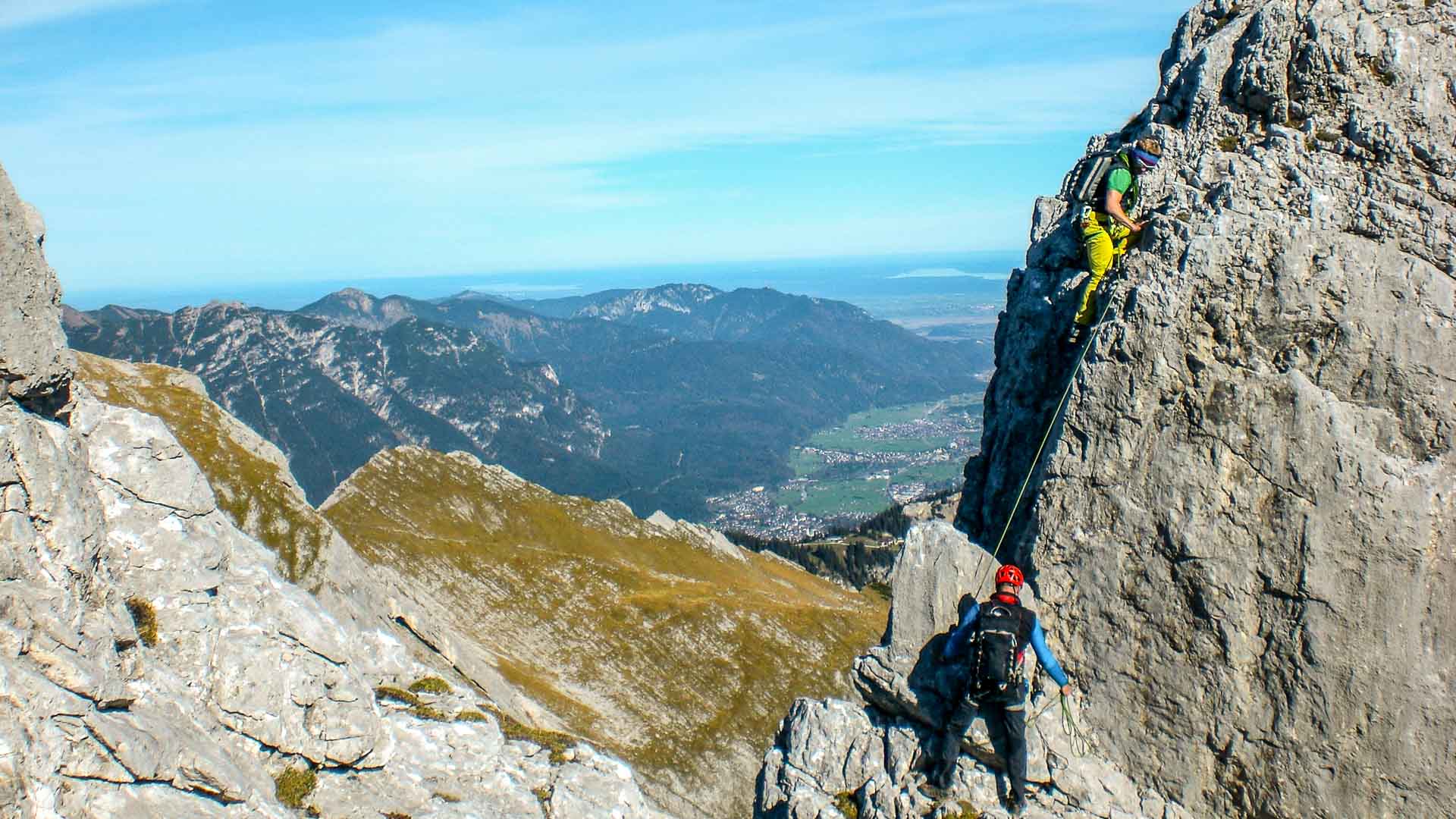 Ein Kletterer sichert am Blassengrat, während ein zweiter die senkrechte Kalkwand mit gelbem Seil aufsteigt.