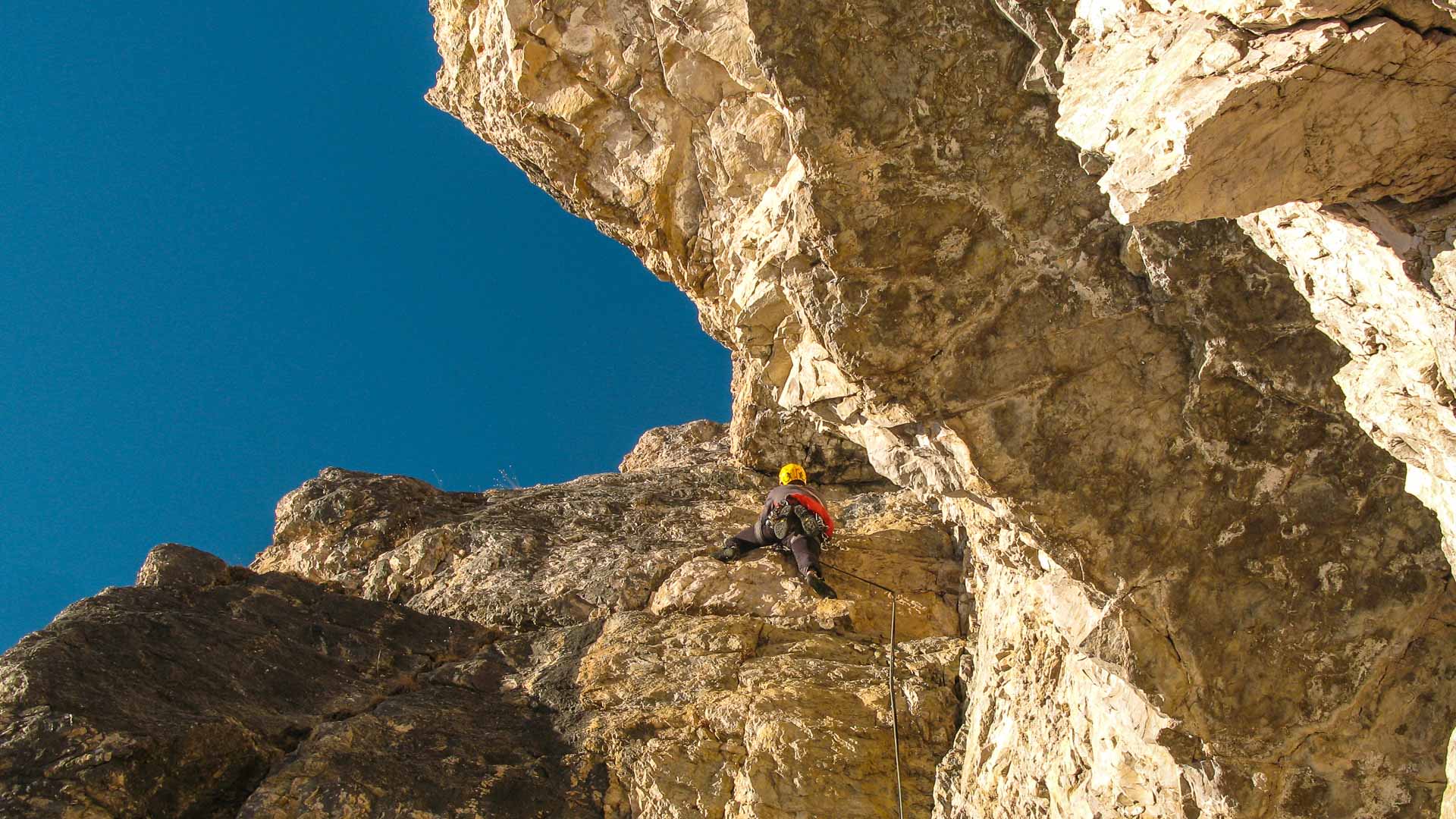 Kletterer mit gelbem Helm und rotem Rucksack an überhängender Kalksteinwand in den Dolomiten, blauer Himmel durch Felsscharte