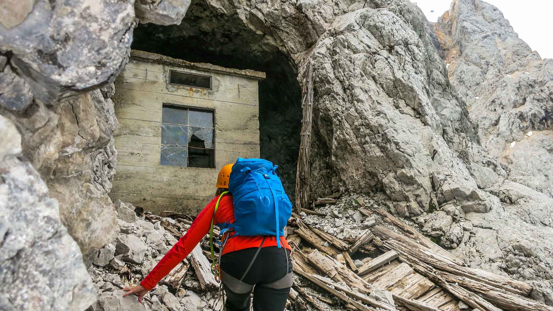 Kletterer mit orangem Helm, rotem Shirt und Klettergurt steigt über Geröll zu einem verfallenen Betongebäude in der Zugspitze