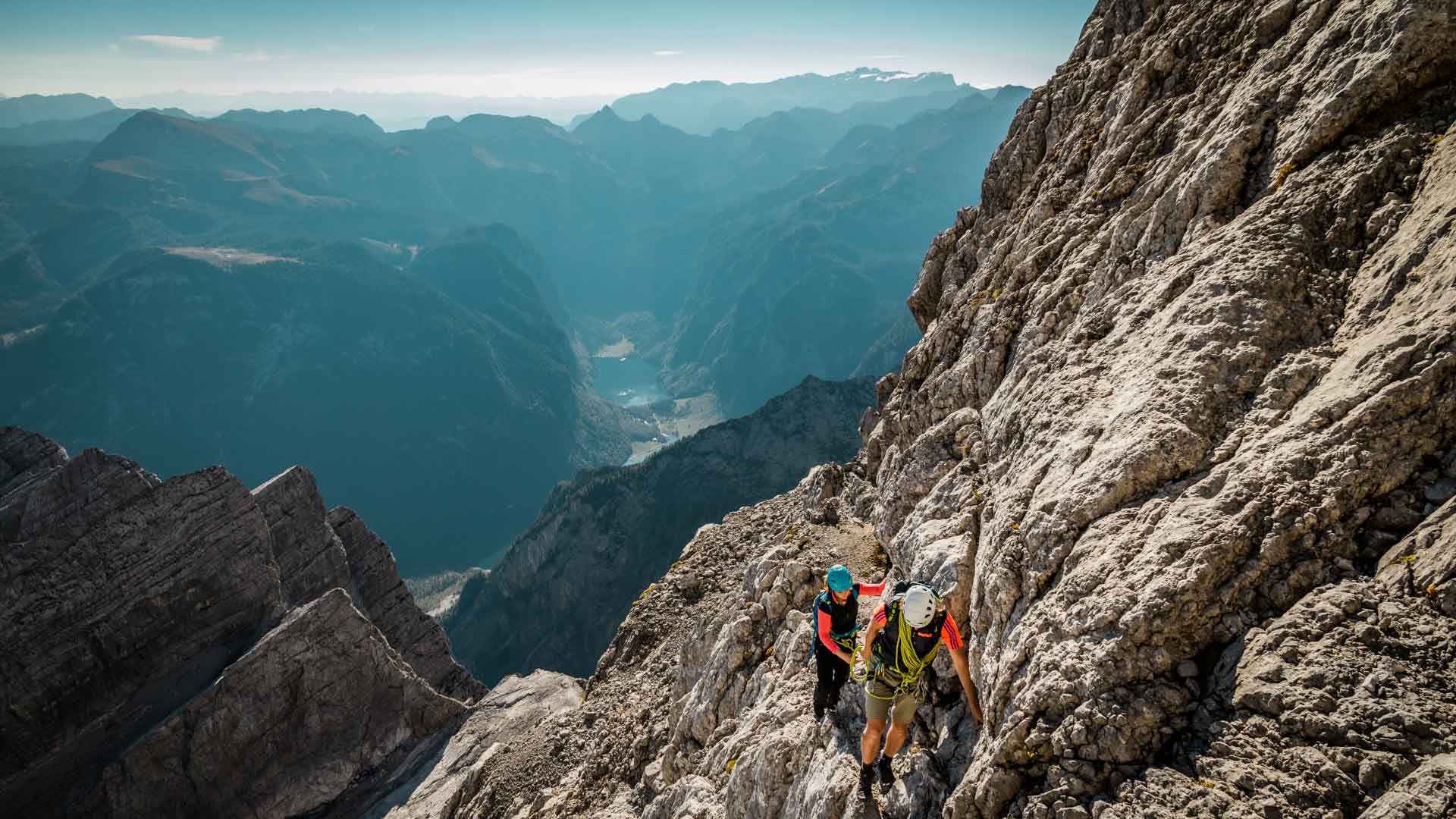 Zwei Kletterer steigen die Kalkflanke der Watzmann-Wiederroute auf, im Hintergrund ein See im Tal und Bergketten.