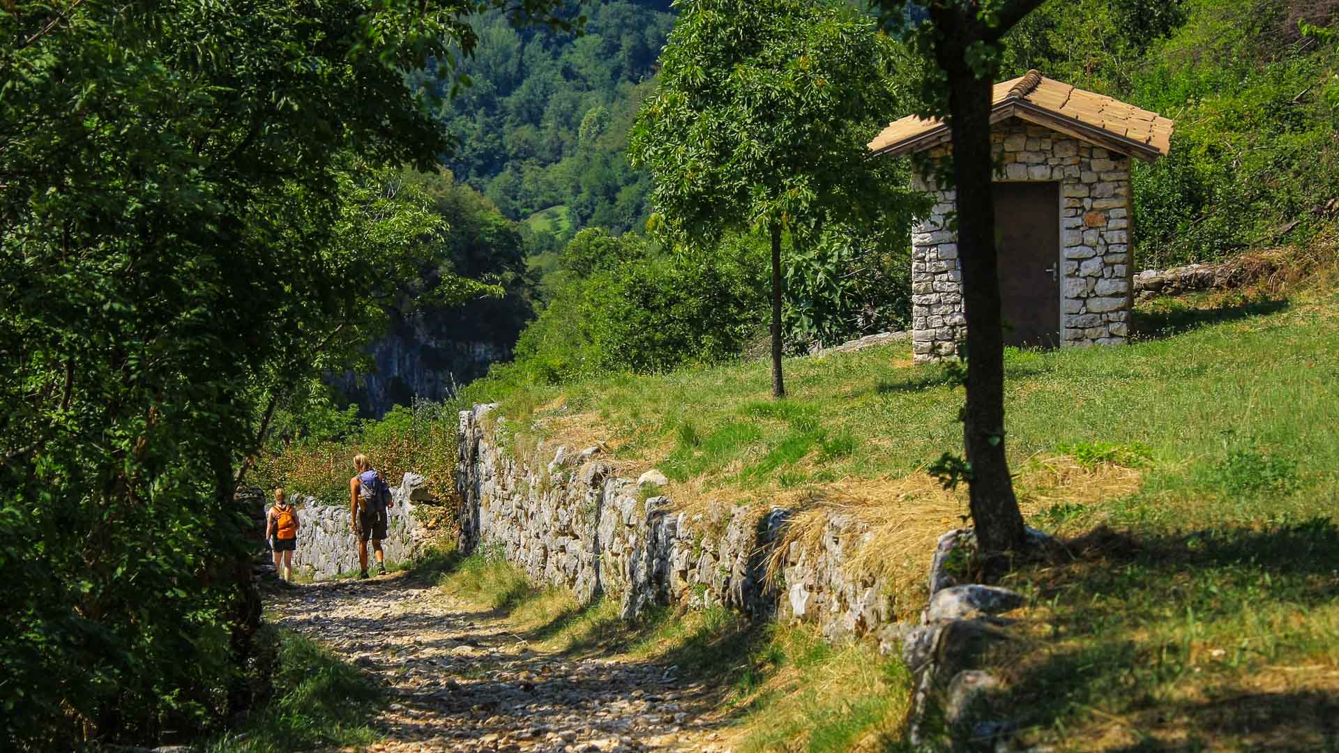 Zwei Kletterer wandern auf gepflastertem Feldweg zwischen Kalksteinmauern in ein bewaldetes Tal am Gardasee.