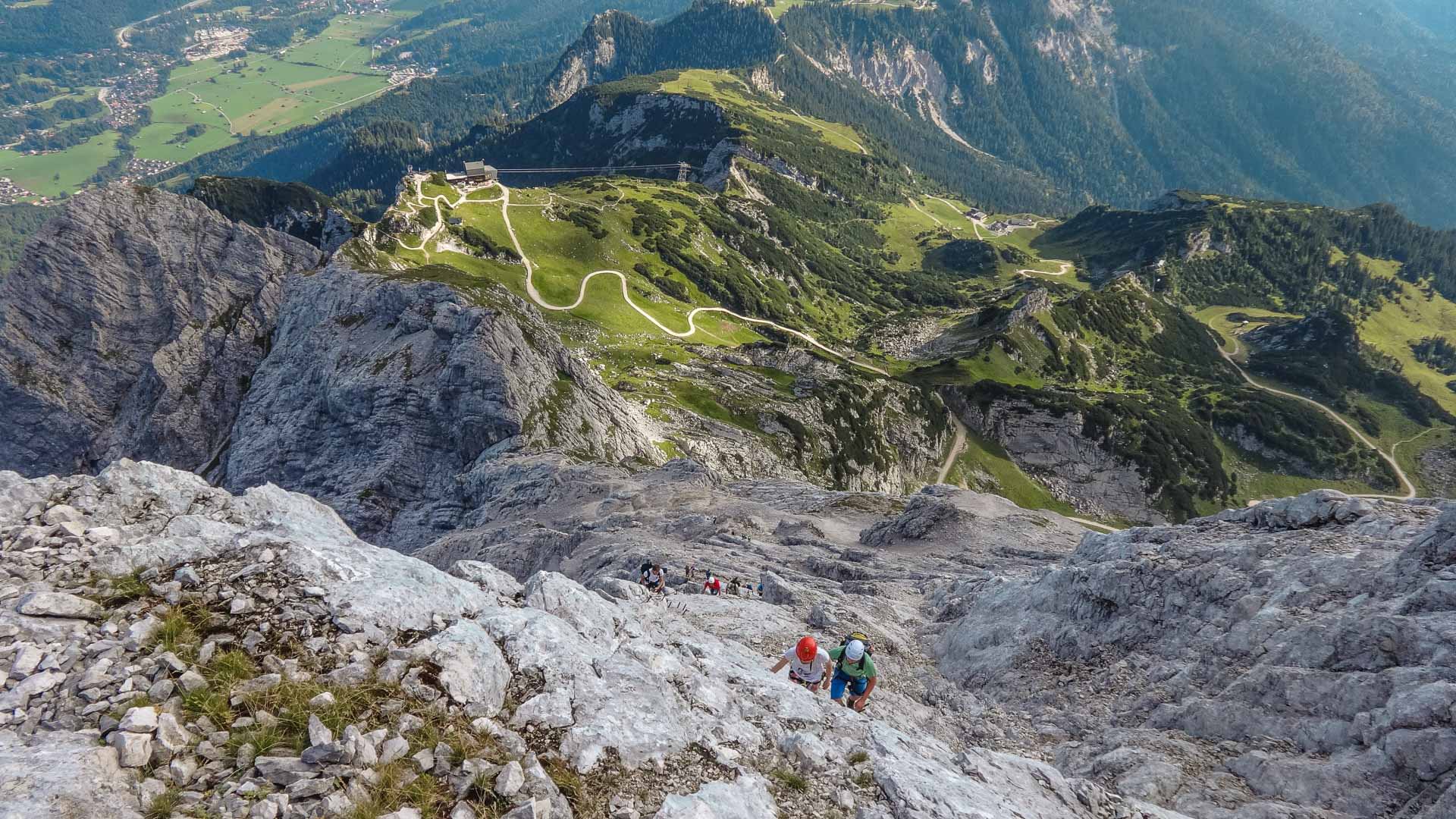 Klettersteiggeher steigen in Vogelperspektive über grauen Kalkfels im Wettersteingebirge auf, im Hintergrund Bergstation