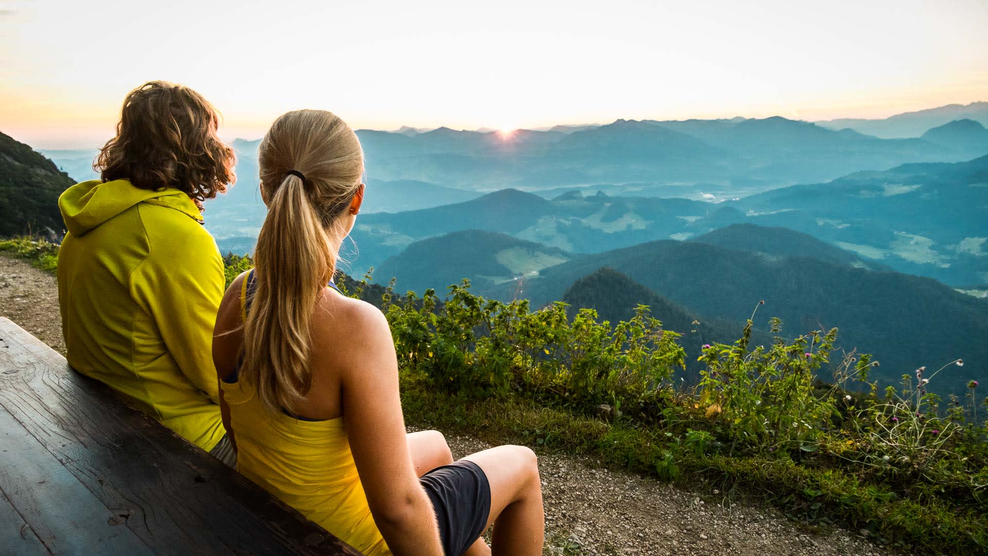 Zwei Klettersteiggeher sitzen auf einer Bank am Scheibenkaser und blicken ins Bergpanorama des Untersberg.