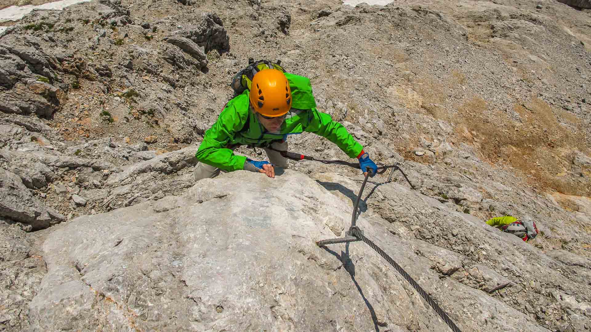 Person mit Petzl-Helm greift am Drahtseil eines Klettersteigs in den Berchtesgadener Alpen eine glatte Kalkfelswand aufwärts