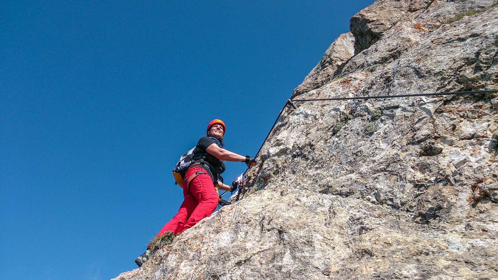 Klettersteiggeher greift am Drahtseil einer hellen Kalkfelswand aufwärts, blauer Himmel im Hintergrund, Dolomiten im Sommer.