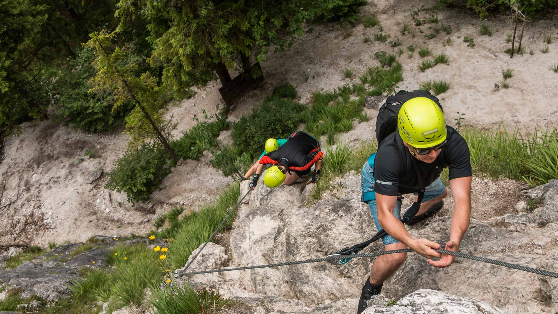 Zwei Klettersteiggeher steigen im Einsteigerkurs bei Berchtesgaden an einem Stahlseil über Kalkfels auf.