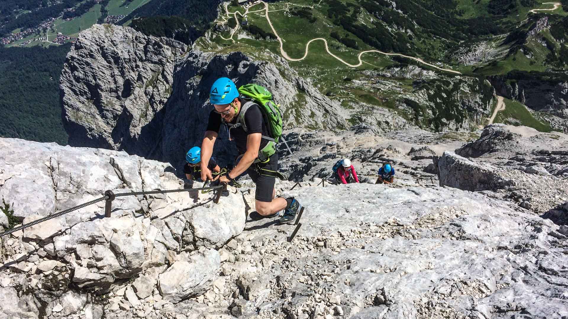 Vier Personen steigen beim Klettersteig-Einsteigerkurs München an Stahlseil und Trittstiften eine helle Kalkfelsplatte