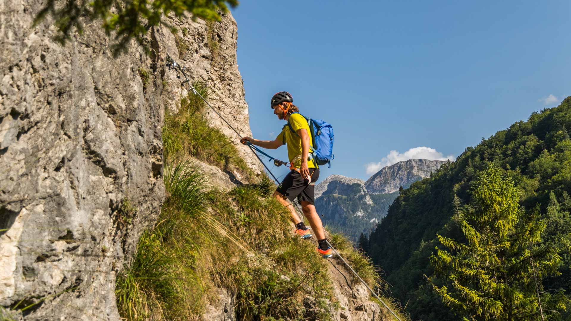 Klettersteiggeher mit Klettersteigset bewegt sich entlang eines grasbedeckten Felsvorsprungs mit Talblick bei Berchtesgaden.