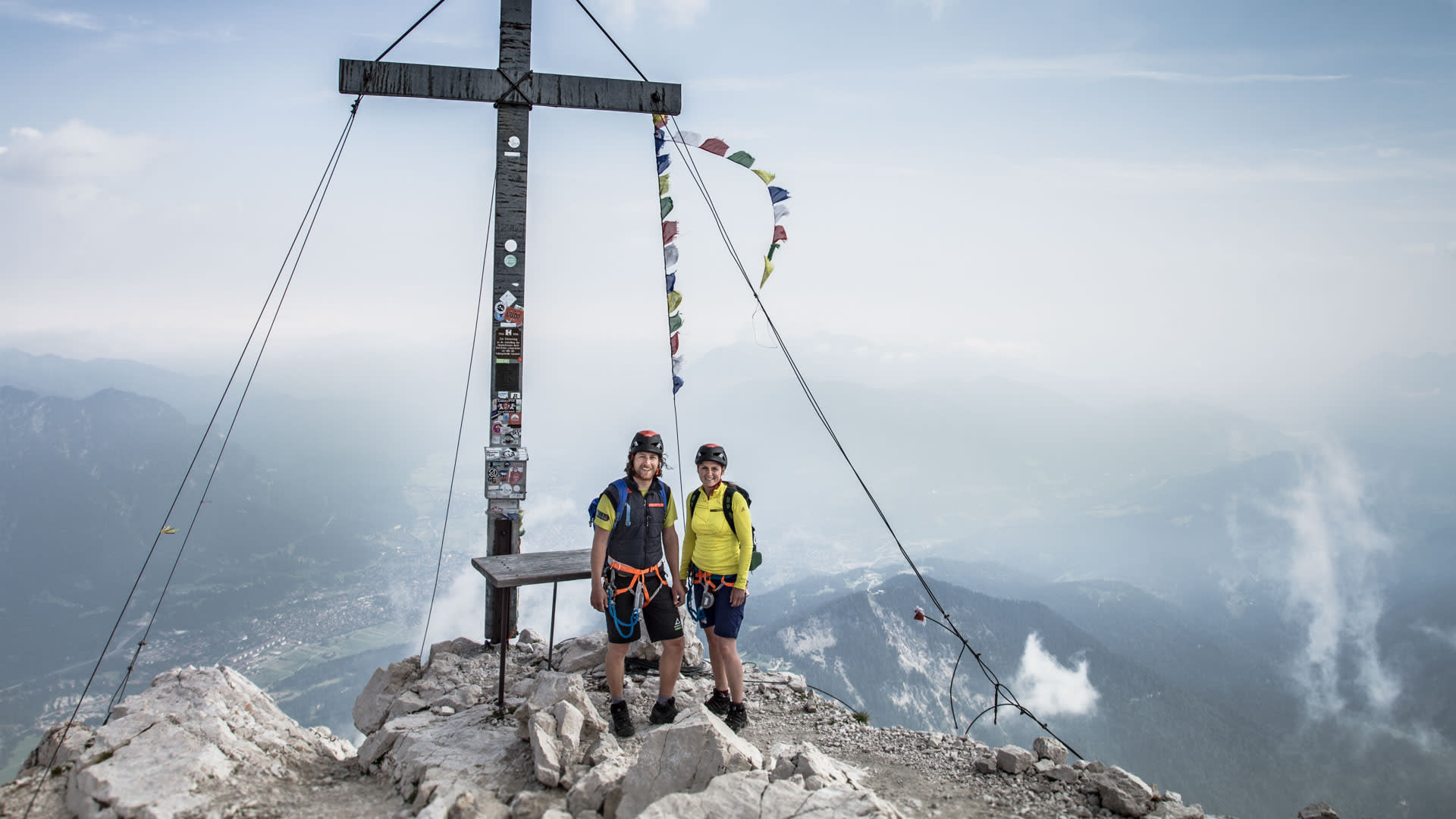 Zwei Klettersteiggeher stehen am Gipfelkreuz der Alpspitze mit Panorama über Garmisch und das Werdenfelser Land.