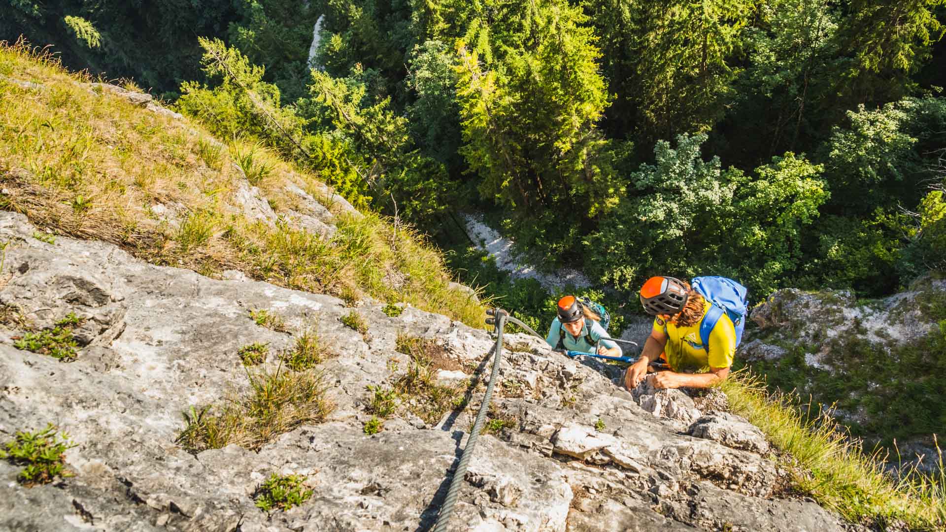 Zwei Klettersteiggeher steigen am Drahtseil des Isidor-Steigs am Grünstein eine Kalkfelswand auf.