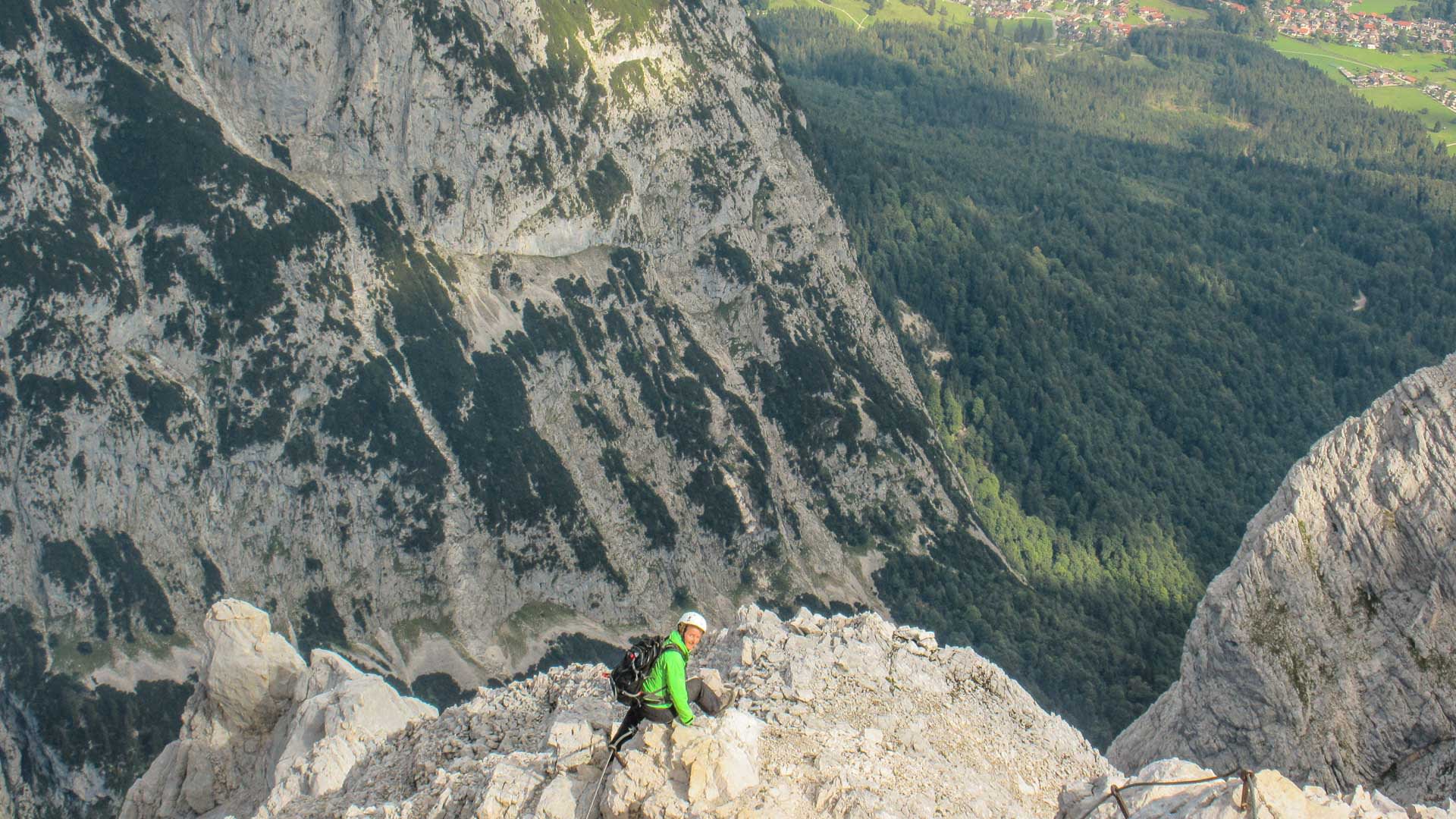 Klettersteiggeher mit grüner Jacke und weißem Helm klettert gesichert auf schmalem Kalksteingrat zur Alpspitze.