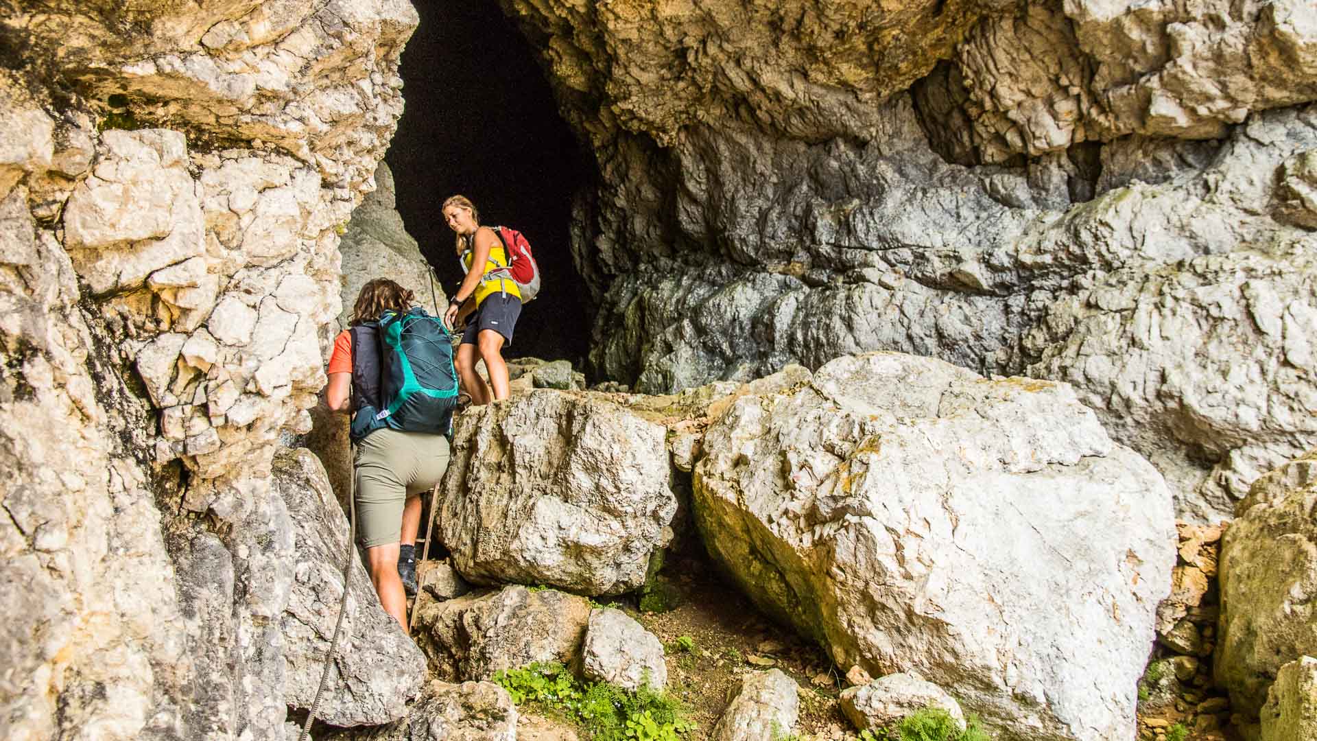 Zwei Klettersteiggeher steigen durch den Felsdurchgang Mittagsloch am Untersberg über Kalksteinblöcke auf.
