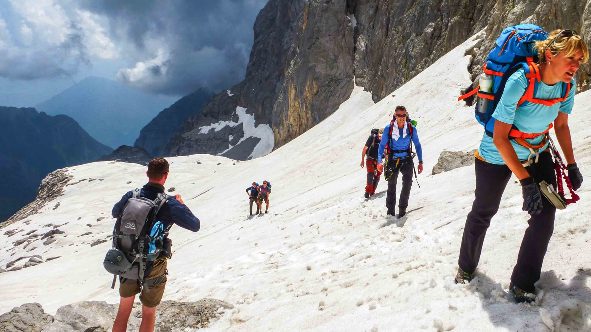 Fünf Klettersteiggeher traversieren einen steilen Sommerschneehang in den Dolomiten der Brenta mit Felskulisse.