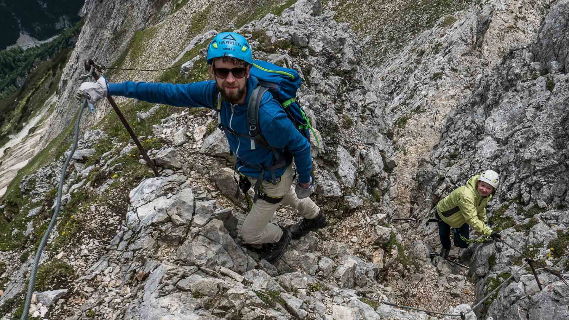 Zwei Klettersteiggeher halten sich an einem Stahlseil an steilem Kalkfels des Mittenwalder Höhenwegs im Karwendel.