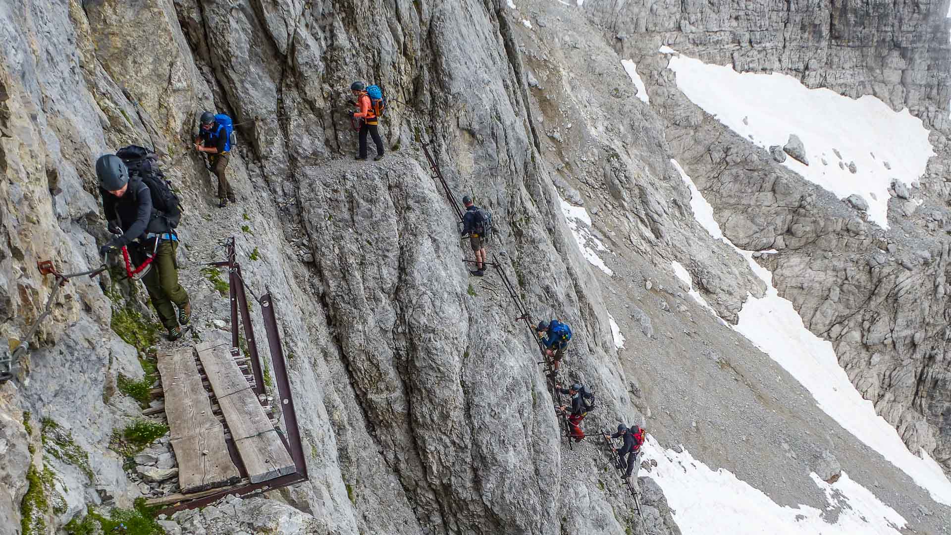 Klettersteig in der Brenta: sechs Klettersteiggeher an Kalkfelswand mit Stahlstiften und Holzsteg-Brücke.
