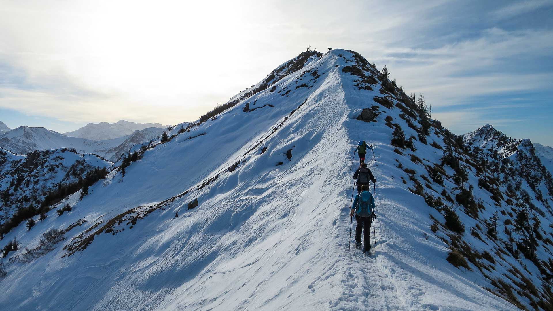 Drei Personen auf Schneeschuhtour steigen hintereinander einen schmalen Schneegratvon Kleinwalsertal zum Gipfelkreuz am Steinmanndl