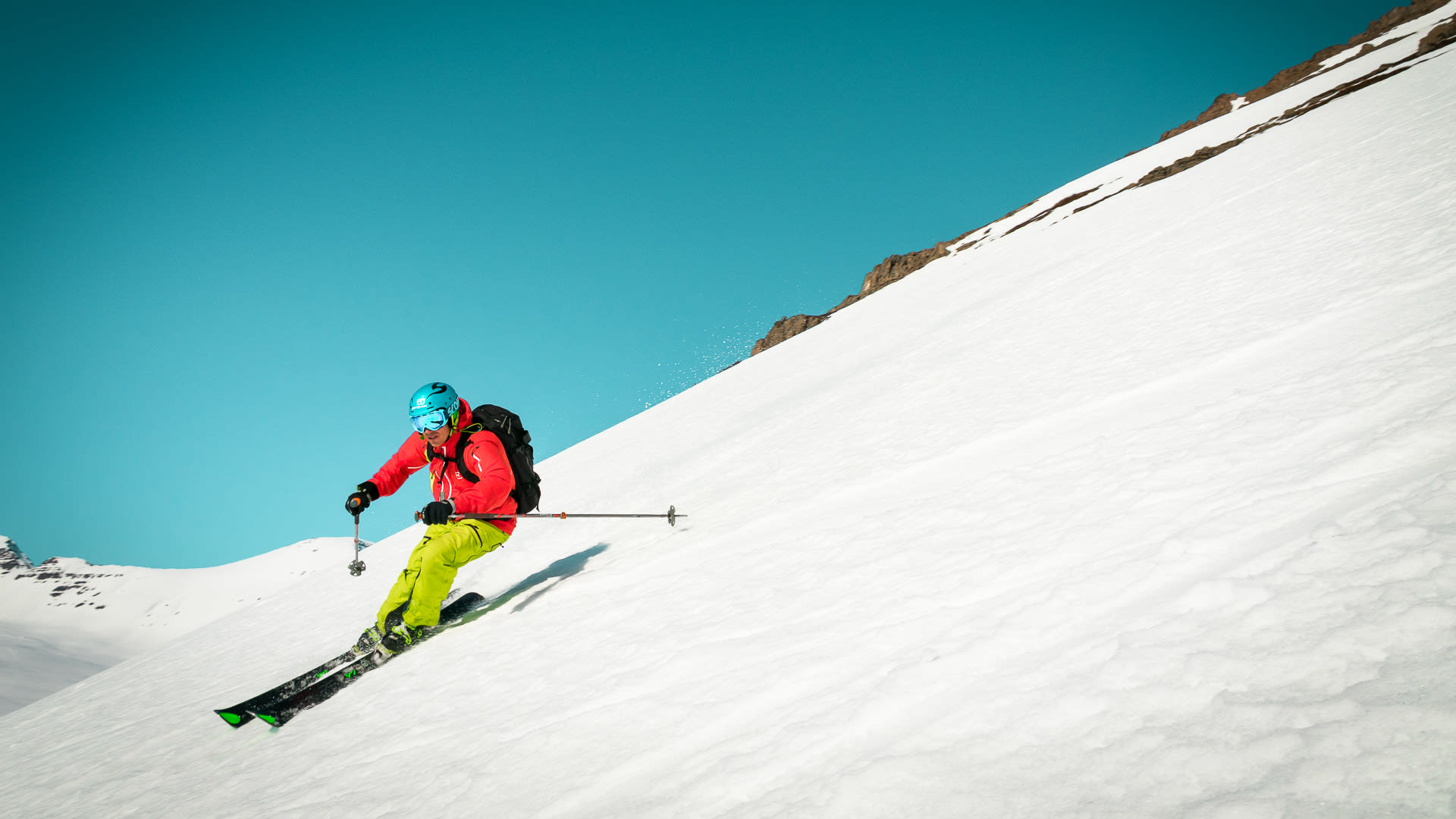 Skitourengeher in roter Jacke fährt steilen Schneehang auf Island im Sonnenschein ab.